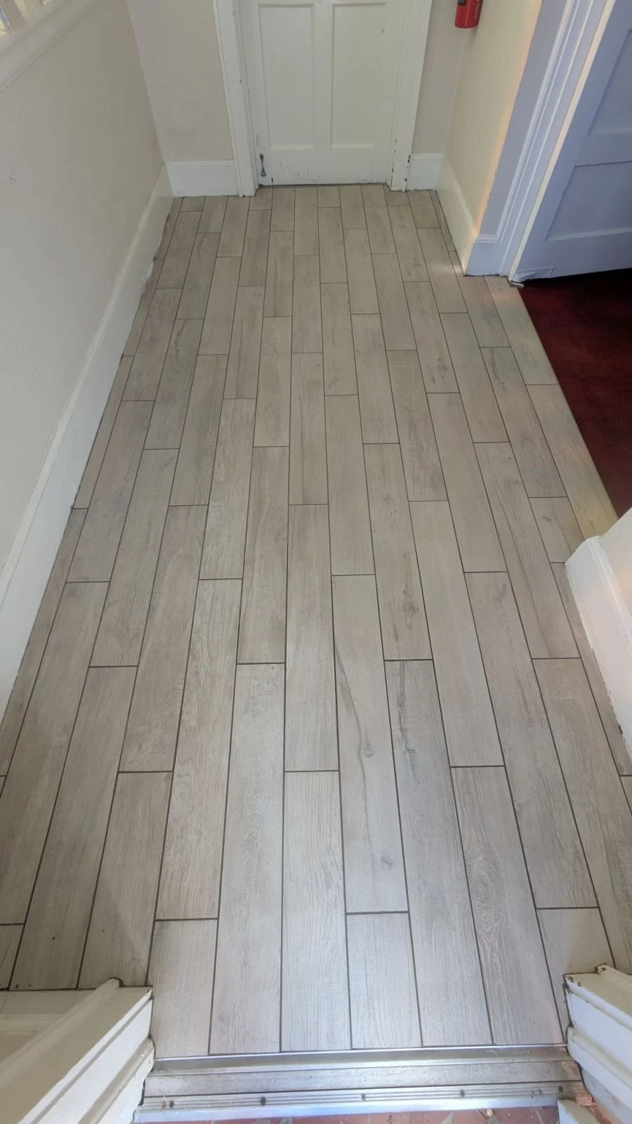 View of a floor with light wood-patterned tiles in a hallway near a closed door, with part of dark wood flooring visible on the right.
