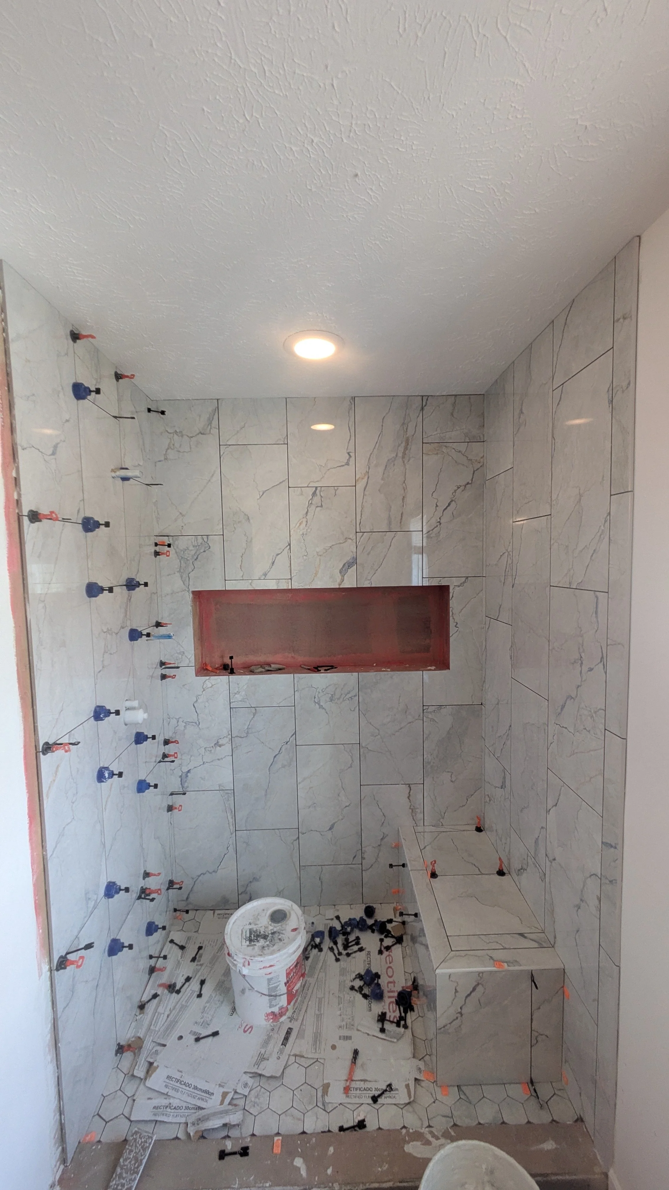 Bathroom under construction with tiles being installed on walls and floor, screws and tools scattered, and a bench.