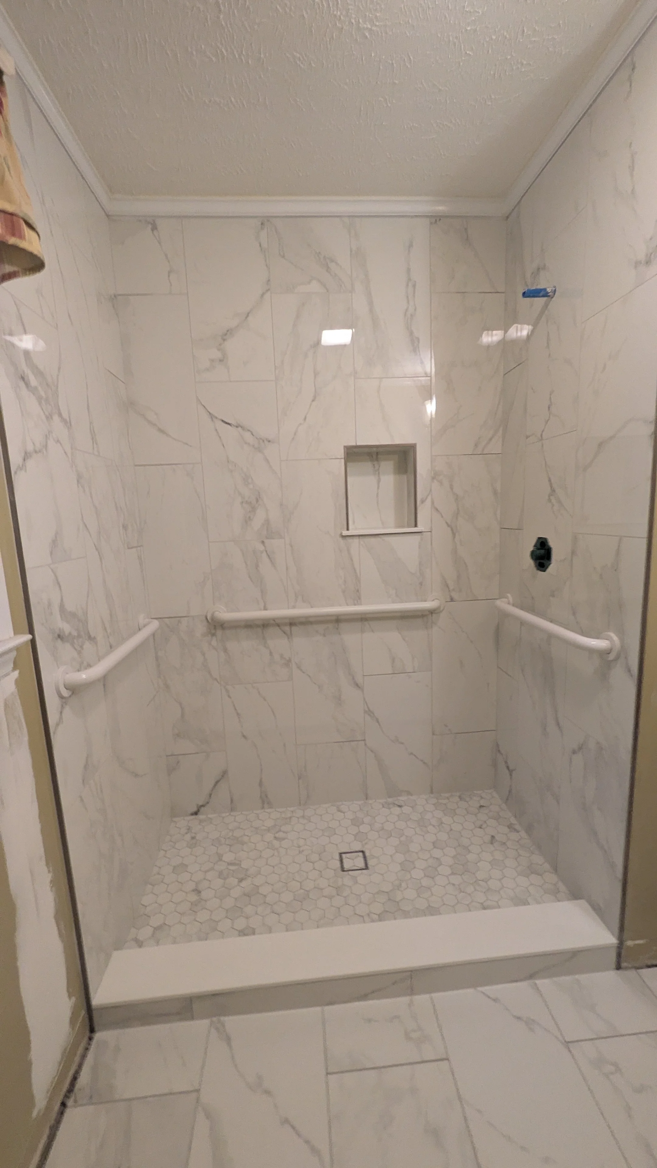 Empty walk-in shower with white marble walls, hexagonal floor tiles, built-in niche, and white grab bars on two sides.