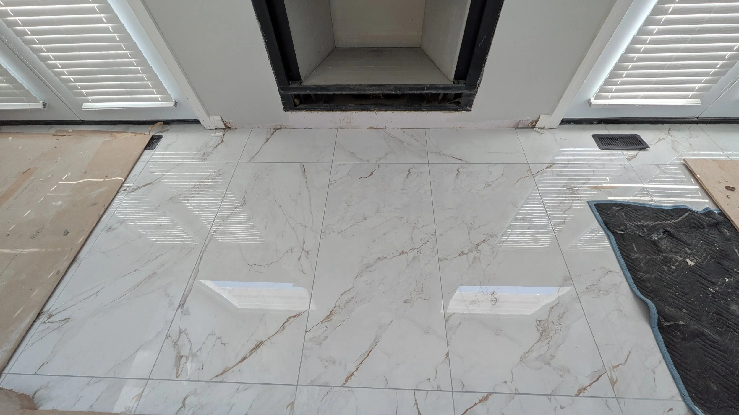 Polished marble floor with two air vents and partially covered area near a door with blinds, and window blinds reflected on the surface.
