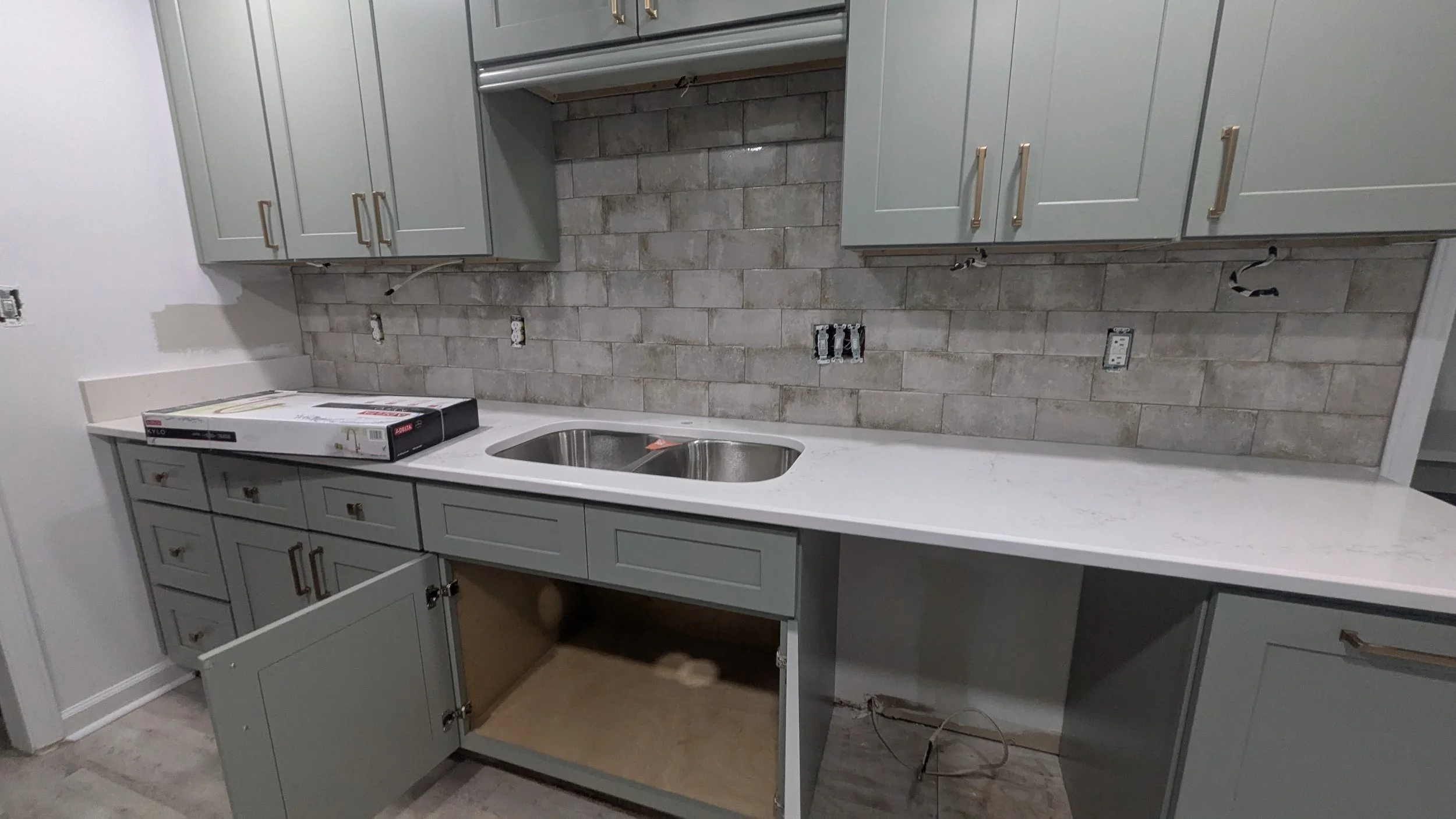 Kitchen in progress with light green cabinets, a white countertop, and a brick-style backsplash wall; some cabinets are open or incomplete, and there are loose wires and tools.
