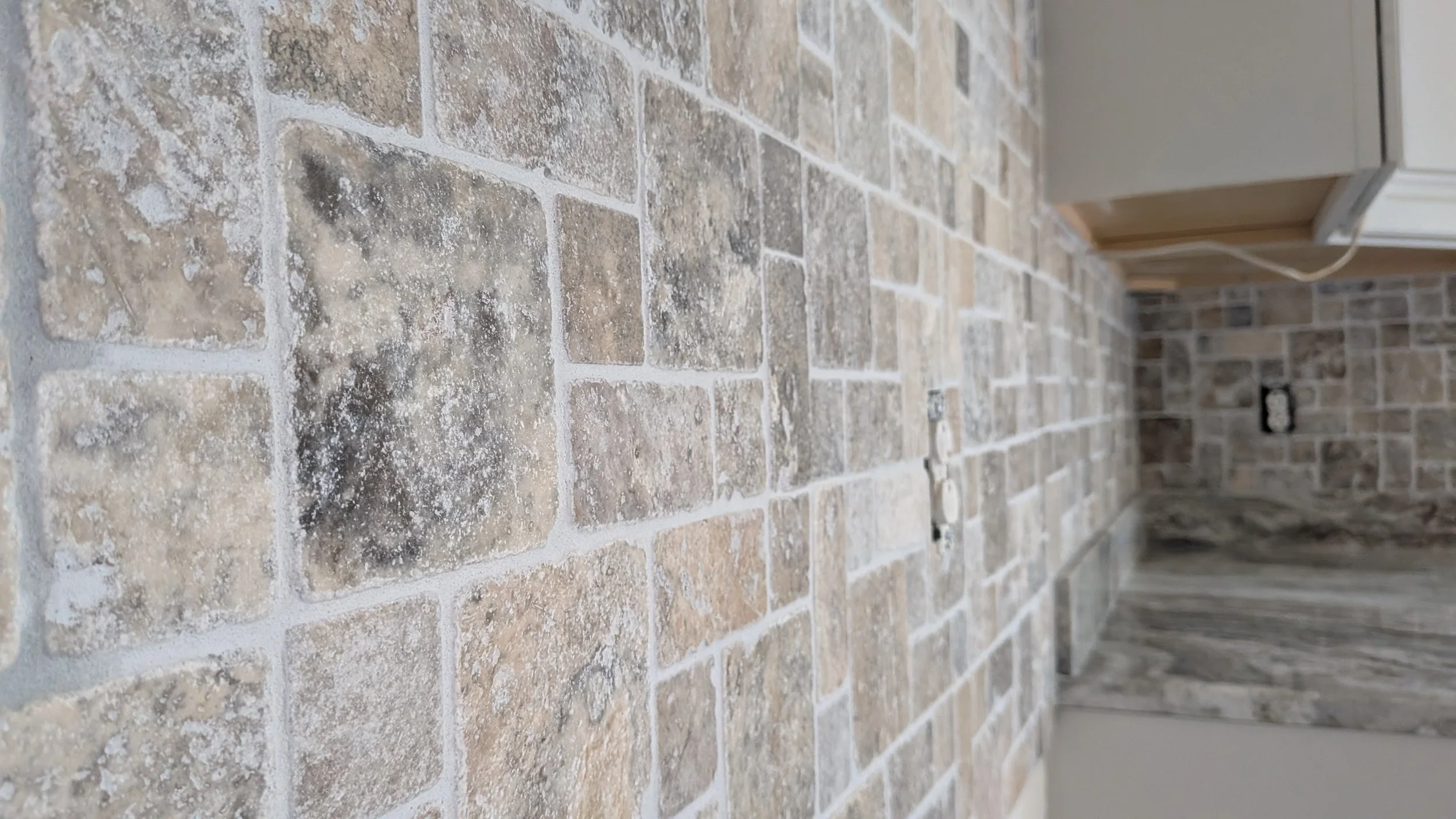 Close-up of a beige and brown stone tile floor in an unfinished room with electrical outlets and wires