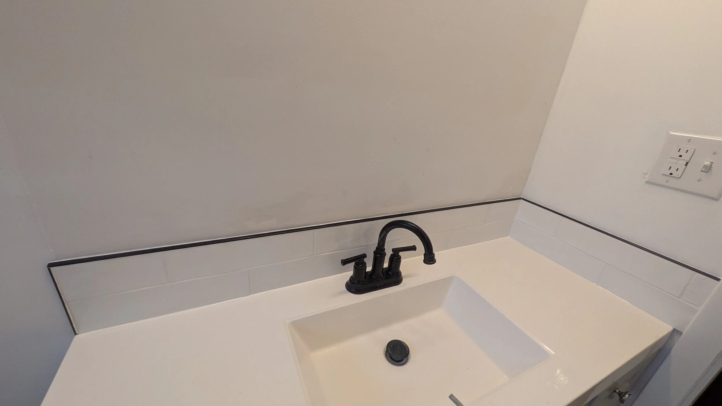 Bathroom vanity with white countertop, black faucet, white tiled backsplash, power outlet on the wall.