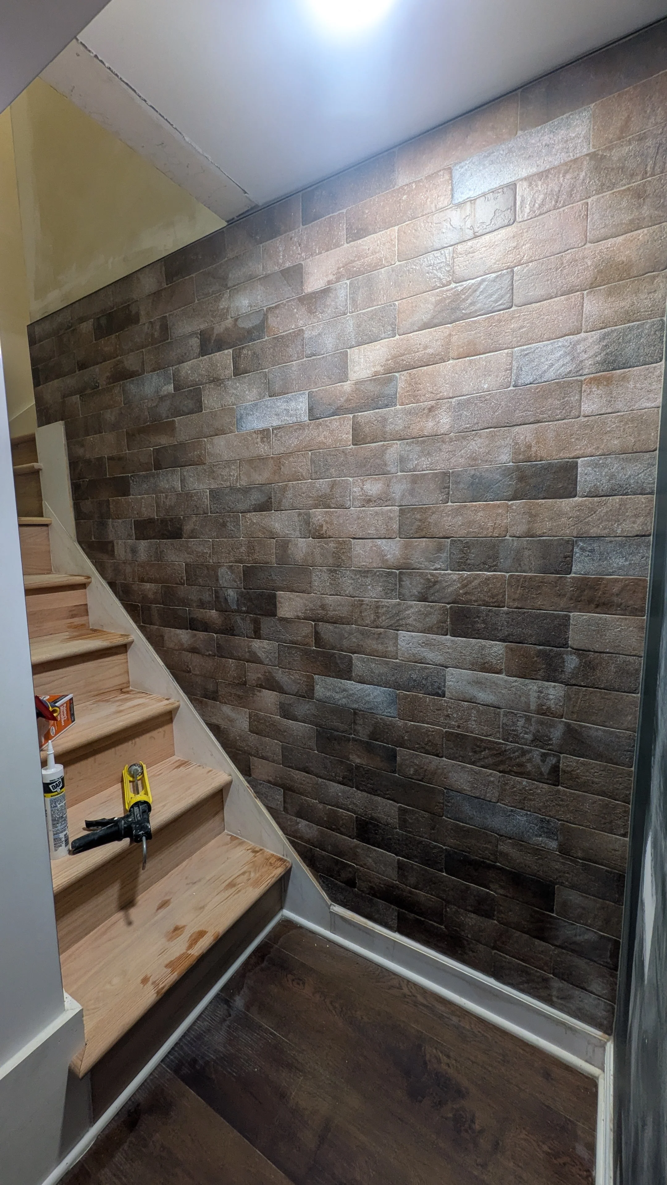 Interior staircase with unfinished wood steps and construction tools, including caulking and a nail gun, next to a brick accent wall in a home renovation project.