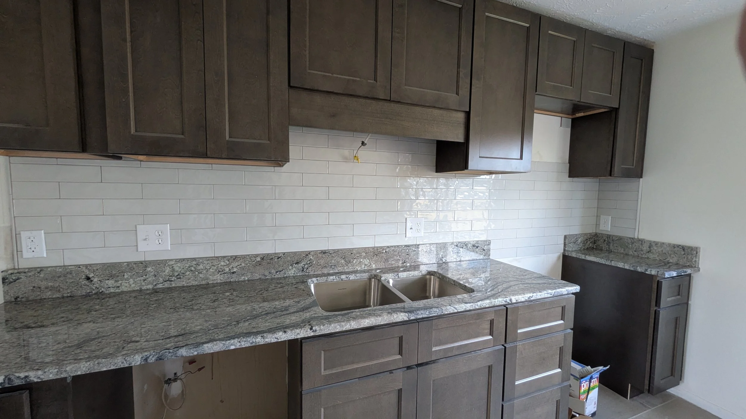 Kitchen with granite countertops, double sink, dark wooden upper and lower cabinets, white subway tile backsplash, and partially visible electrical outlets.