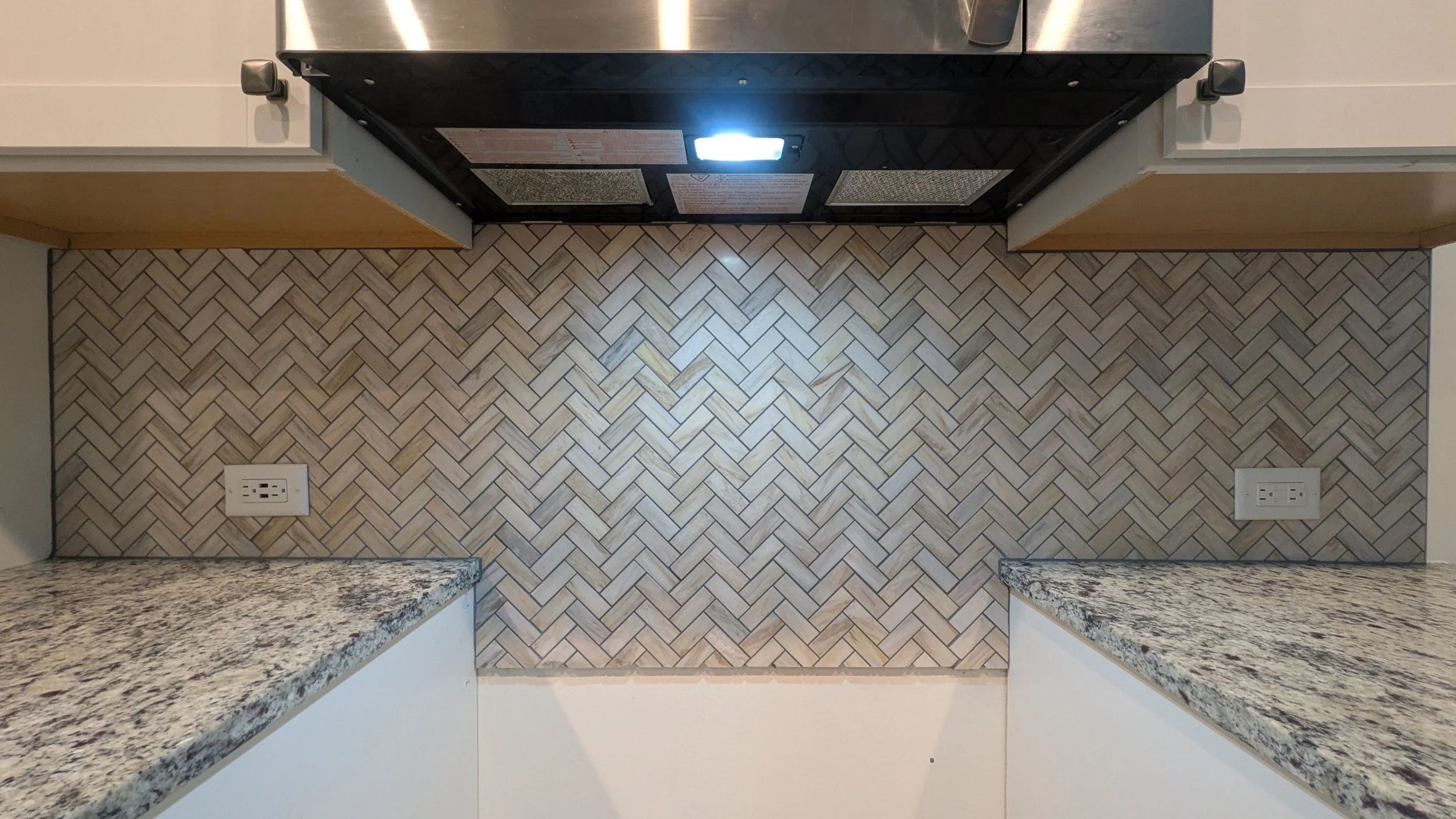 View of a kitchen wall with a black stove in the center, two electrical outlets on each side, granite countertops at the bottom, and beige herringbone-patterned tile backsplash.