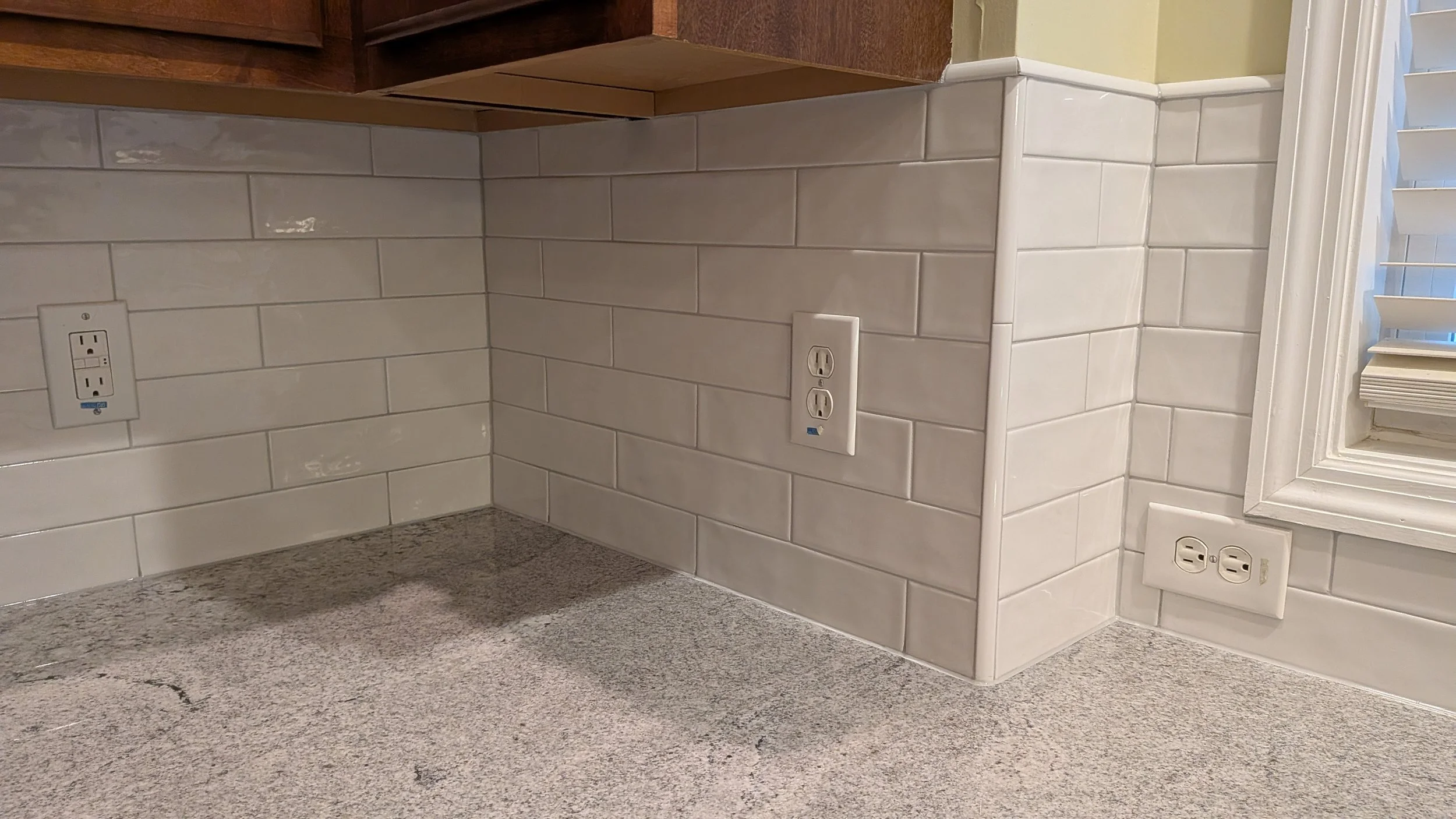 Corner of a kitchen countertop with three electrical outlets on the walls, white subway tiles as backsplash, beige carpeted floor, and a window with blinds.