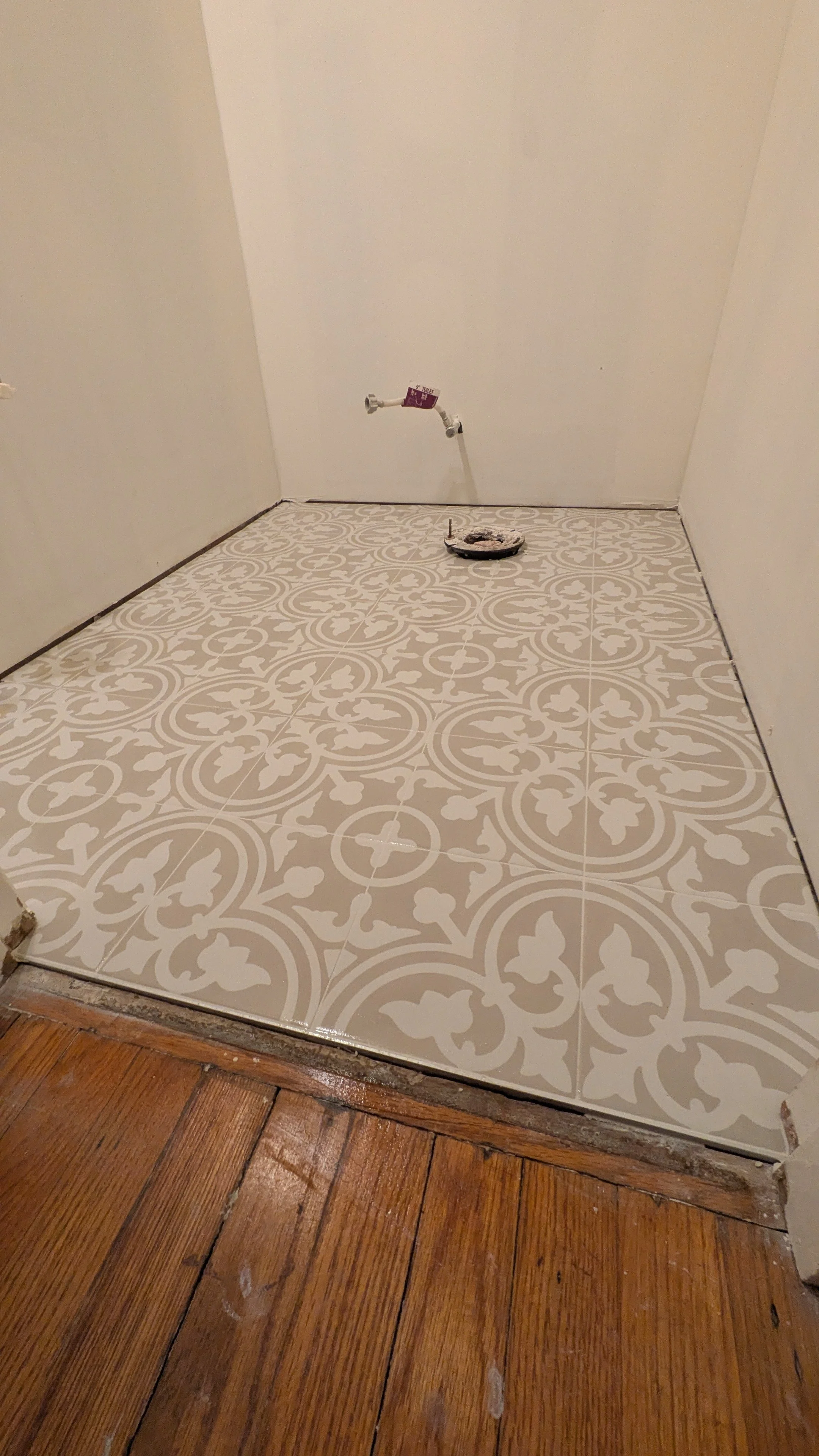 Room with new patterned floor tiles and hardwood flooring, plumbing pipe on white wall, and a round drain cover on the tiles.