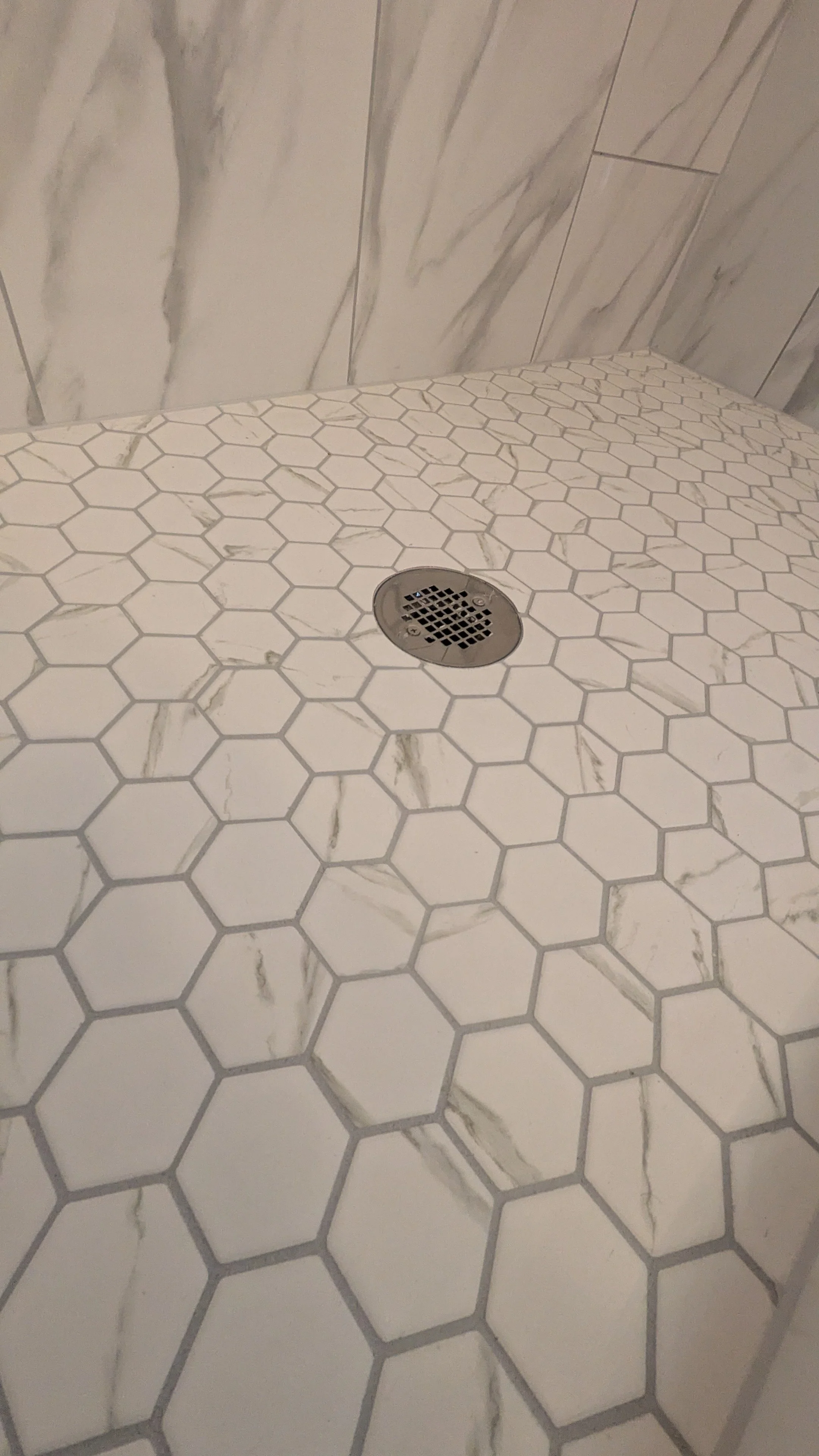 Close-up of a tiled shower floor with white hexagonal tiles and gray grout, with a round metal drain in the center, and marble tiles on the wall in the background.