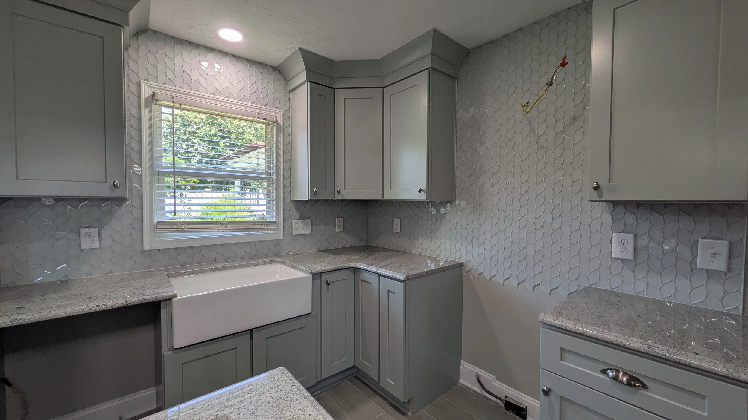Kitchen with light gray cabinets and granite countertops, white farmhouse sink under window, textured leaf-patterned backsplash, and multiple electrical outlets, with some stray wiring visible on the wall.