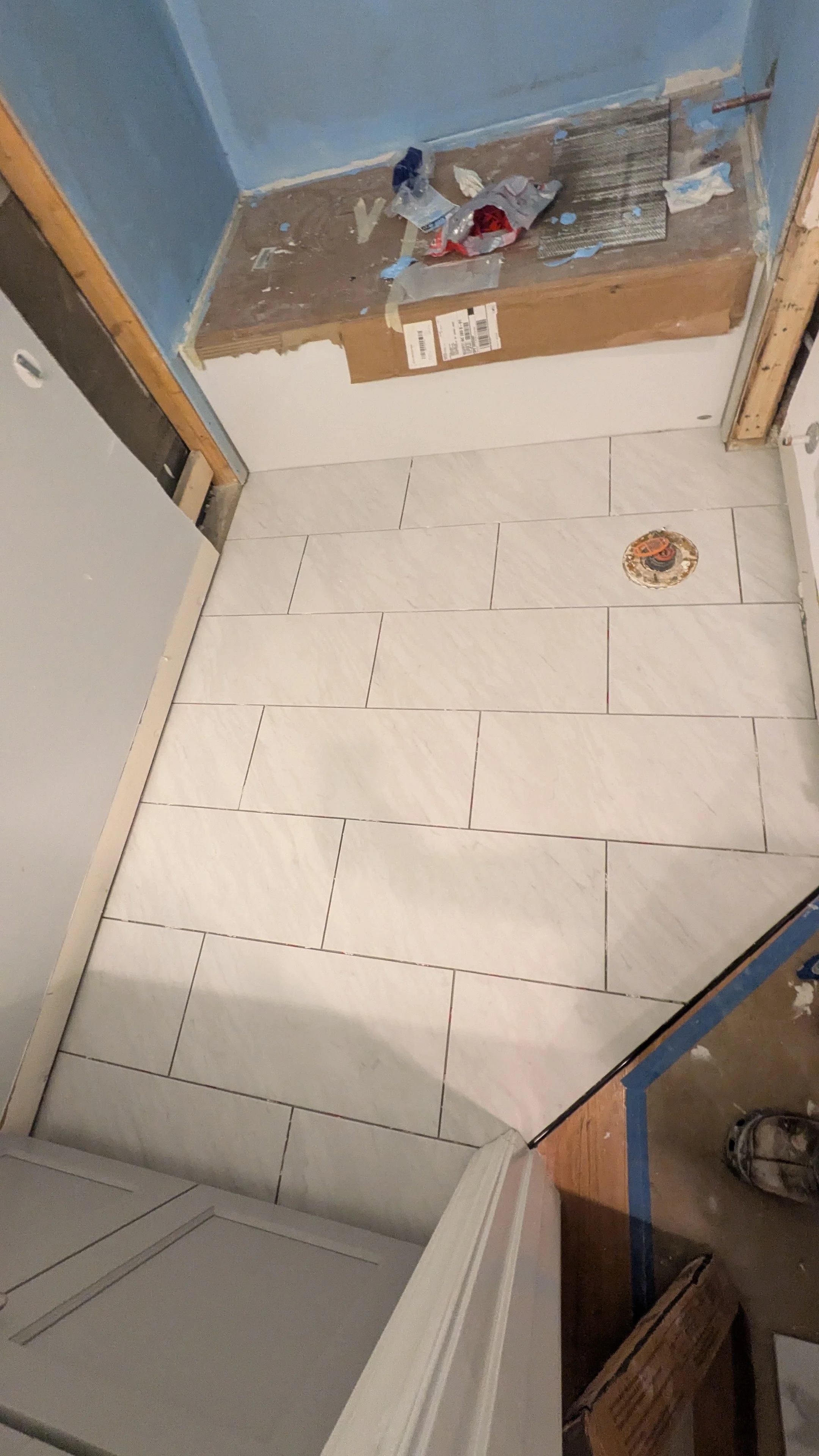 A room undergoing renovation with newly installed white ceramic floor tiles, a partially constructed doorway with unfinished trim, and construction materials on a small raised platform against a blue wall.