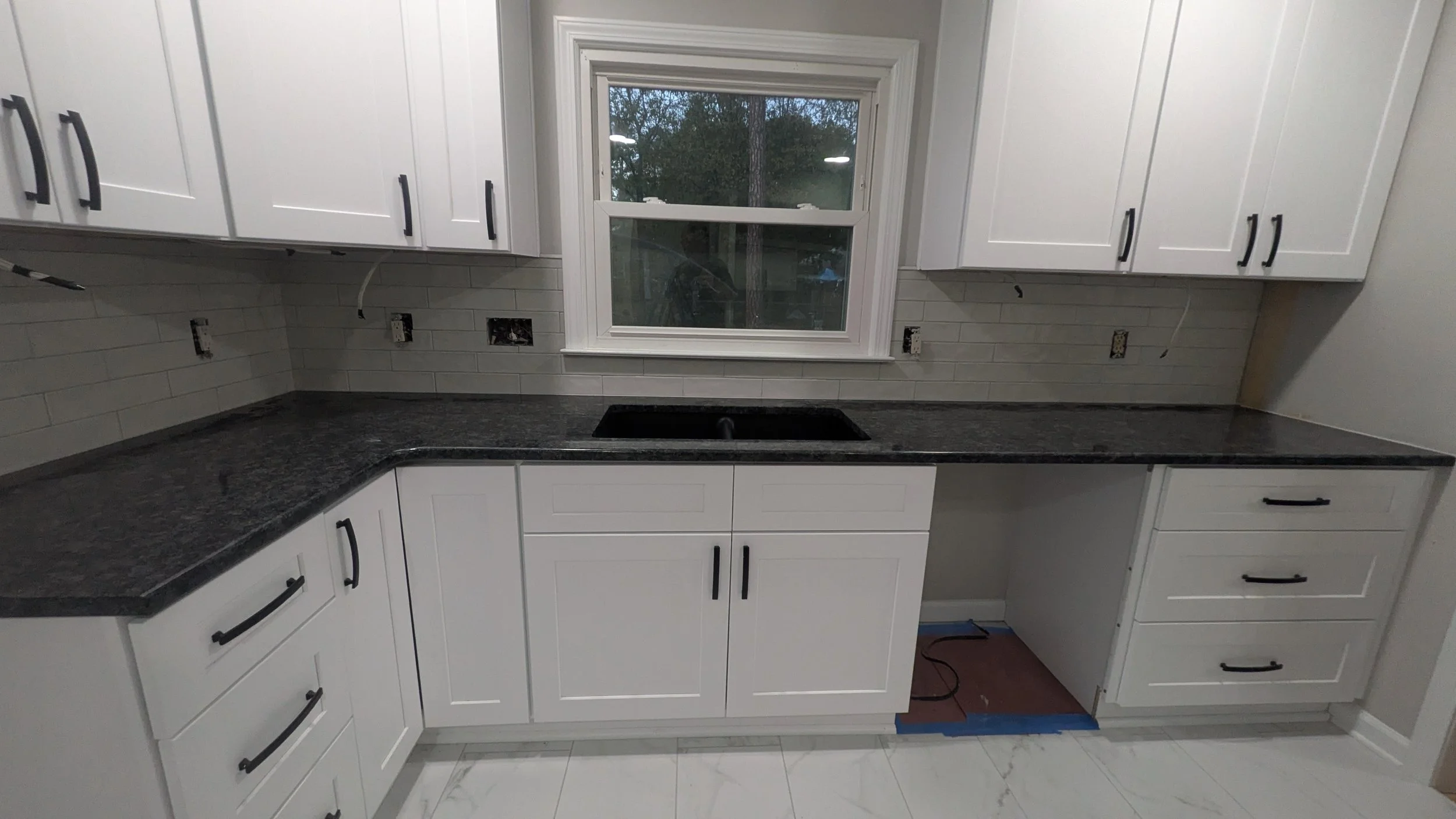 Kitchen with white cabinets, black handles, black countertop, window above, and a black sink installed. Wall with tile backsplash and multiple electrical outlets, some with open wiring. The floor is white with gray veining.