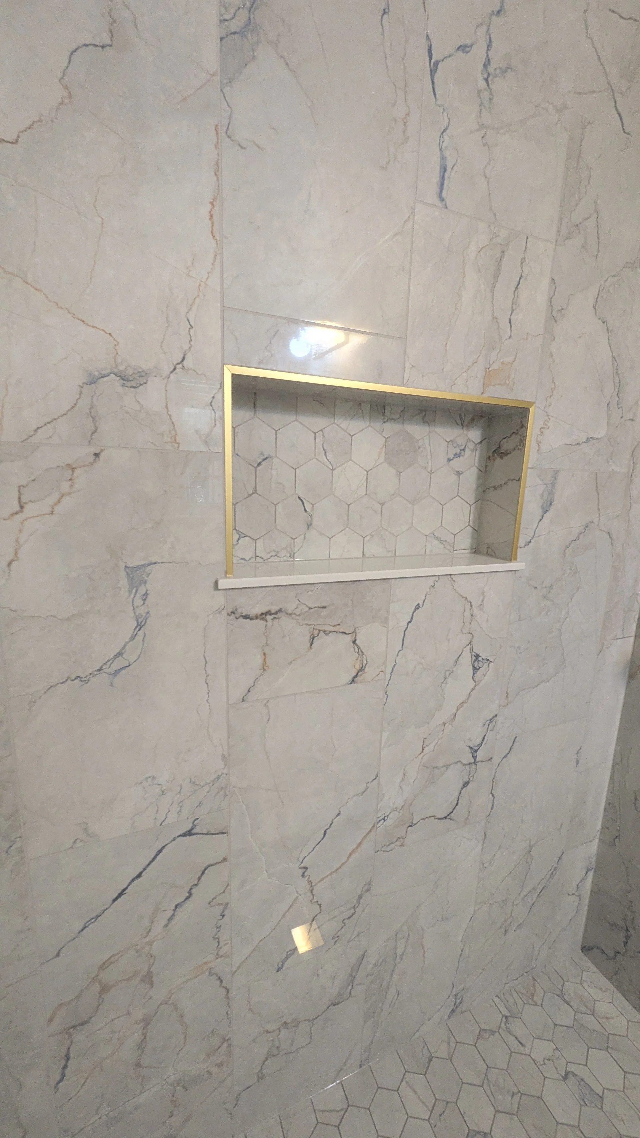 Close-up of a shower wall with large marble tiles, featuring a built-in hexagonal tile niche accented with a gold border and a small white ledge below.