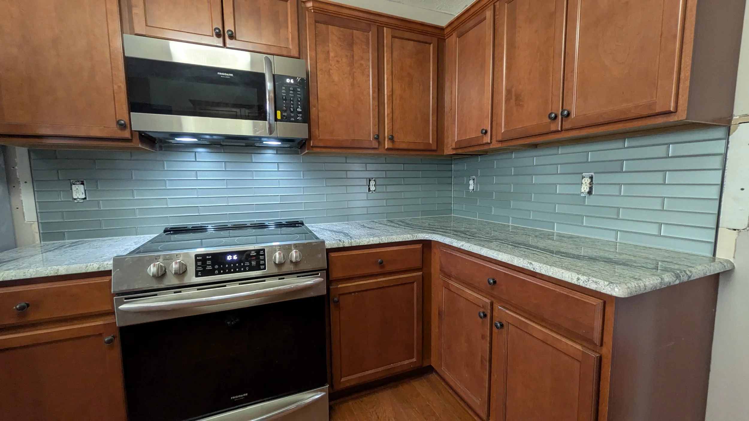Kitchen with wooden cabinets, granite countertops, a gray tile backsplash, a microwave oven, and a stainless steel stove.