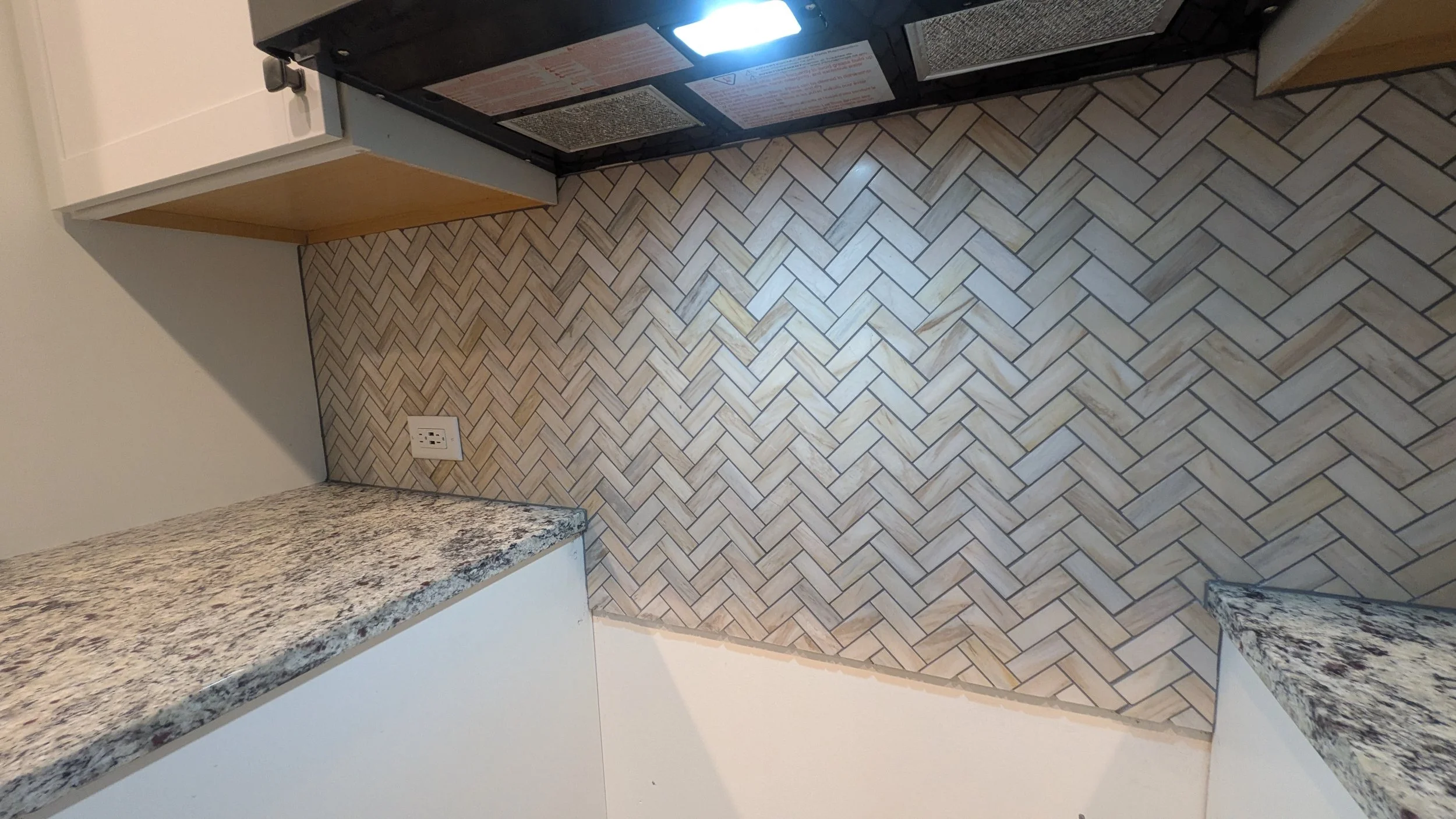 A kitchen corner with a black microwave, granite countertops, a white wall with a power outlet, and a herringbone-patterned tile floor.