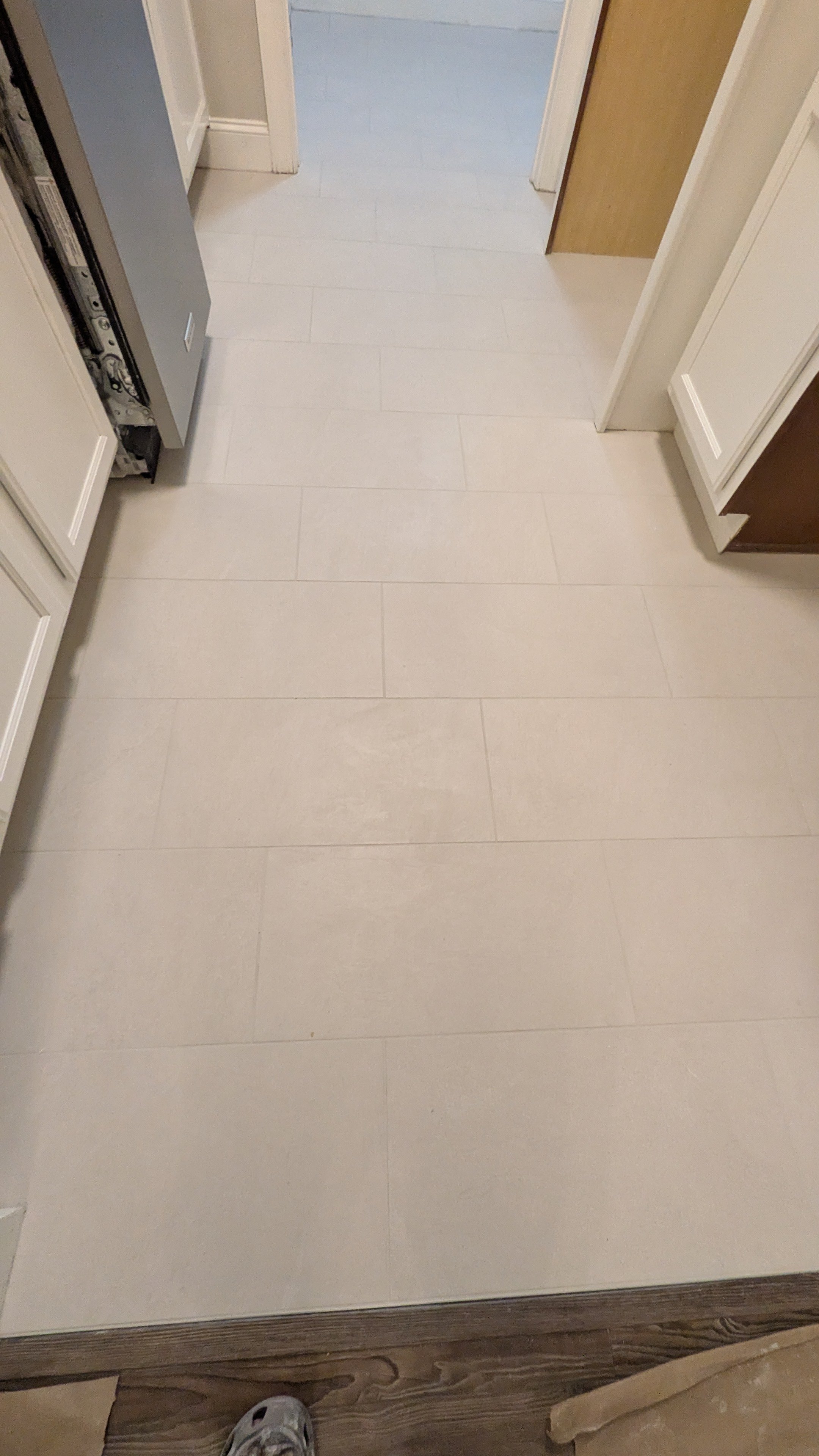 Interior view of a kitchen with beige tiled flooring, white cabinetry, and a part of a refrigerator on the left side. An open doorway leads to a room with blue flooring and a wooden door.