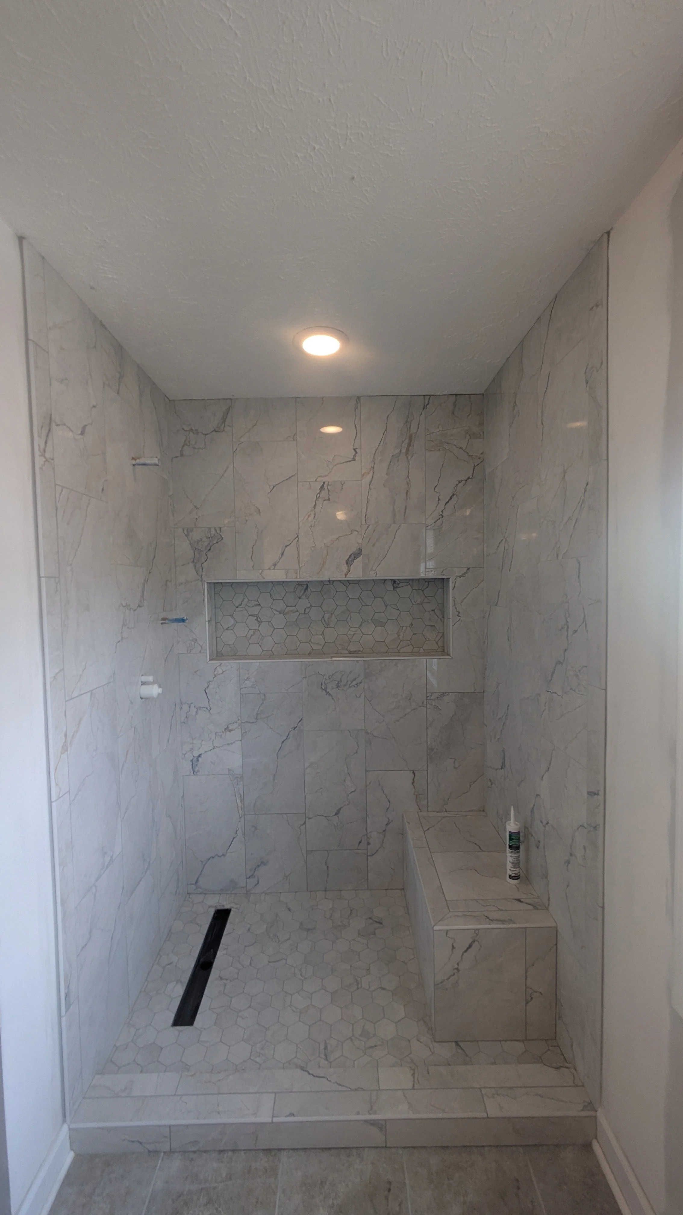 A newly constructed walk-in shower with marble tiles, a built-in bench, a recessed niche with hexagonal tiles, and a linear drain on the floor.
