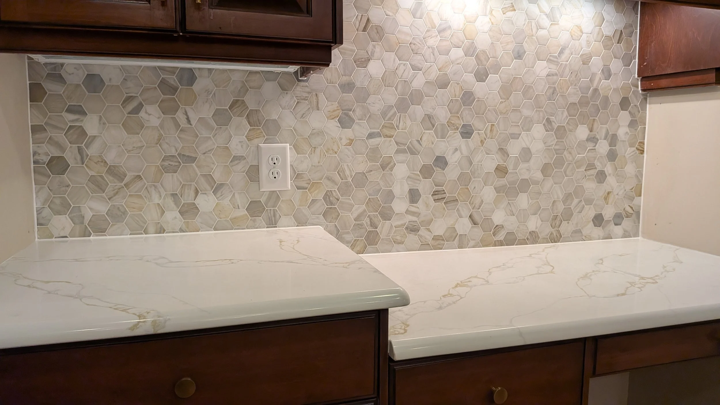 Kitchen countertop with marble pattern, dark wooden cabinets above, and a hexagon tile backsplash in shades of gray and beige, with an electrical outlet on the wall.