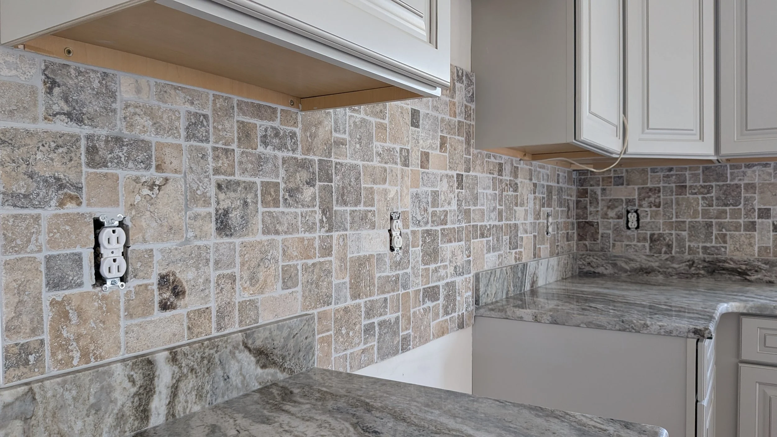 Kitchen backsplash with beige and brown stone tiles, empty wall outlets, granite countertop, and white kitchen cabinets.