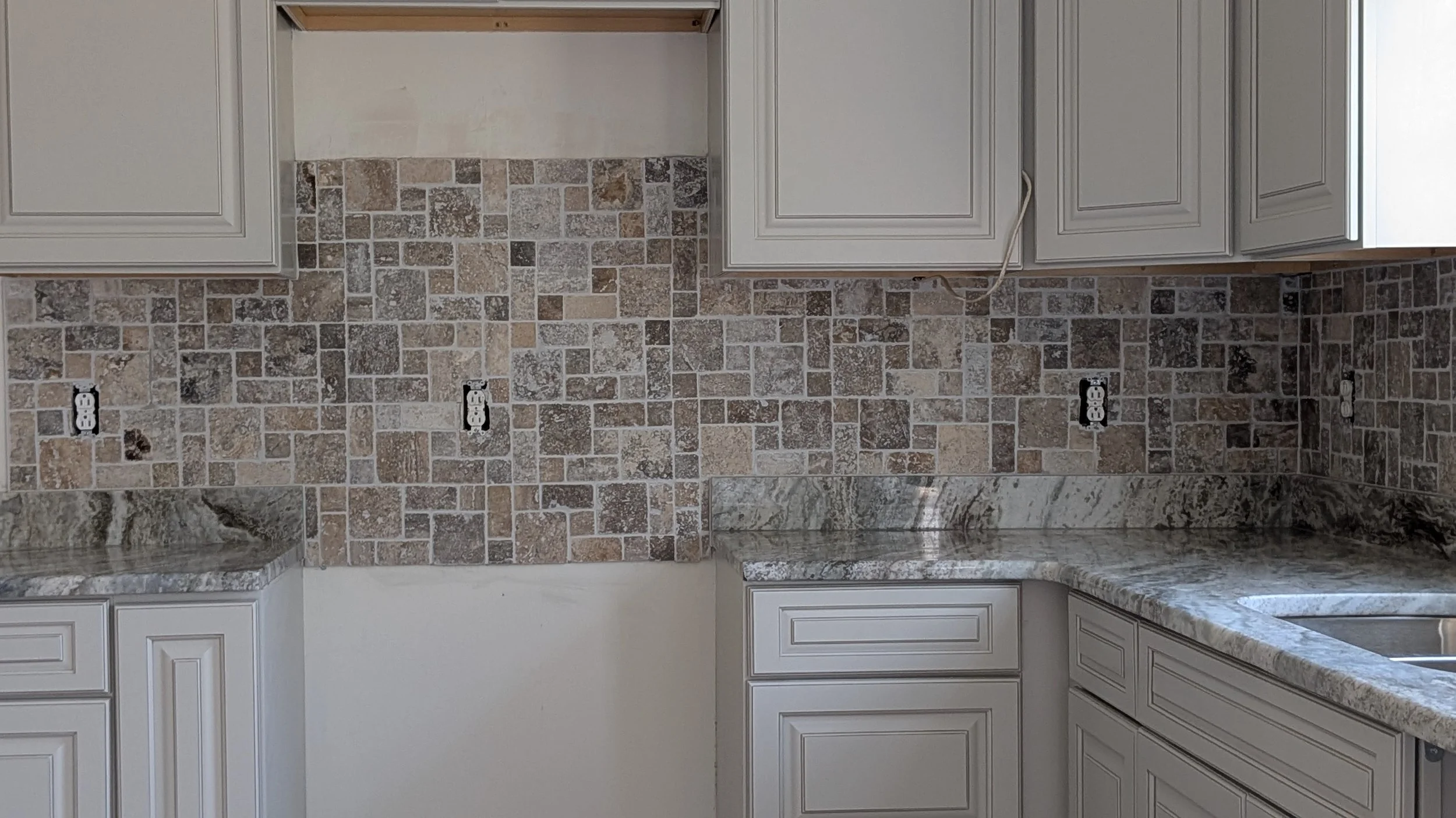 Kitchen countertop with granite surface, beige cabinets, and a tile backsplash in shades of brown and gray. There are four electrical outlets on the backsplash.