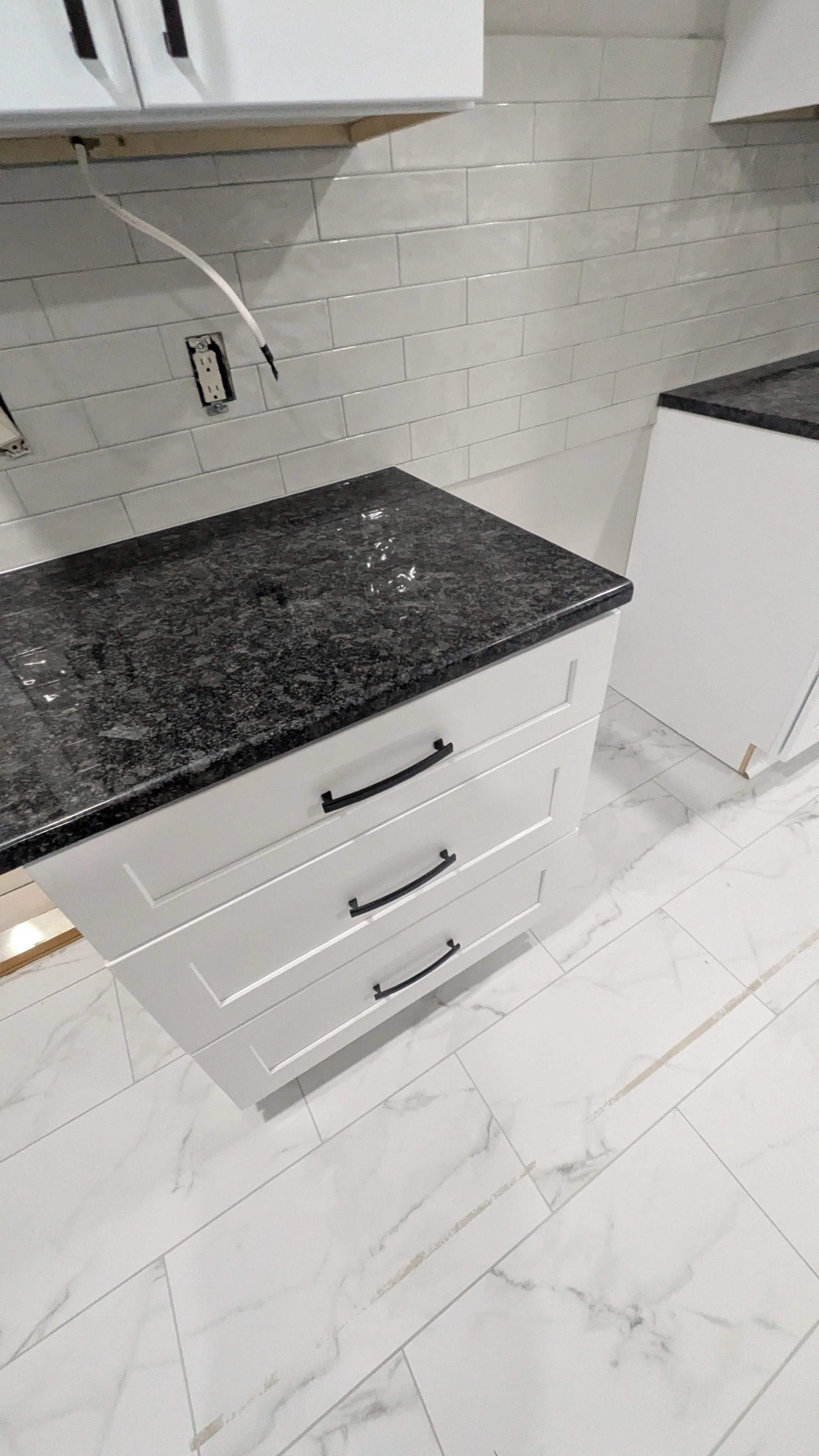 Empty kitchen with white cabinets, black granite countertops, and a white tiled wall. There are faucet and electrical outlets, one with a wire hanging out.