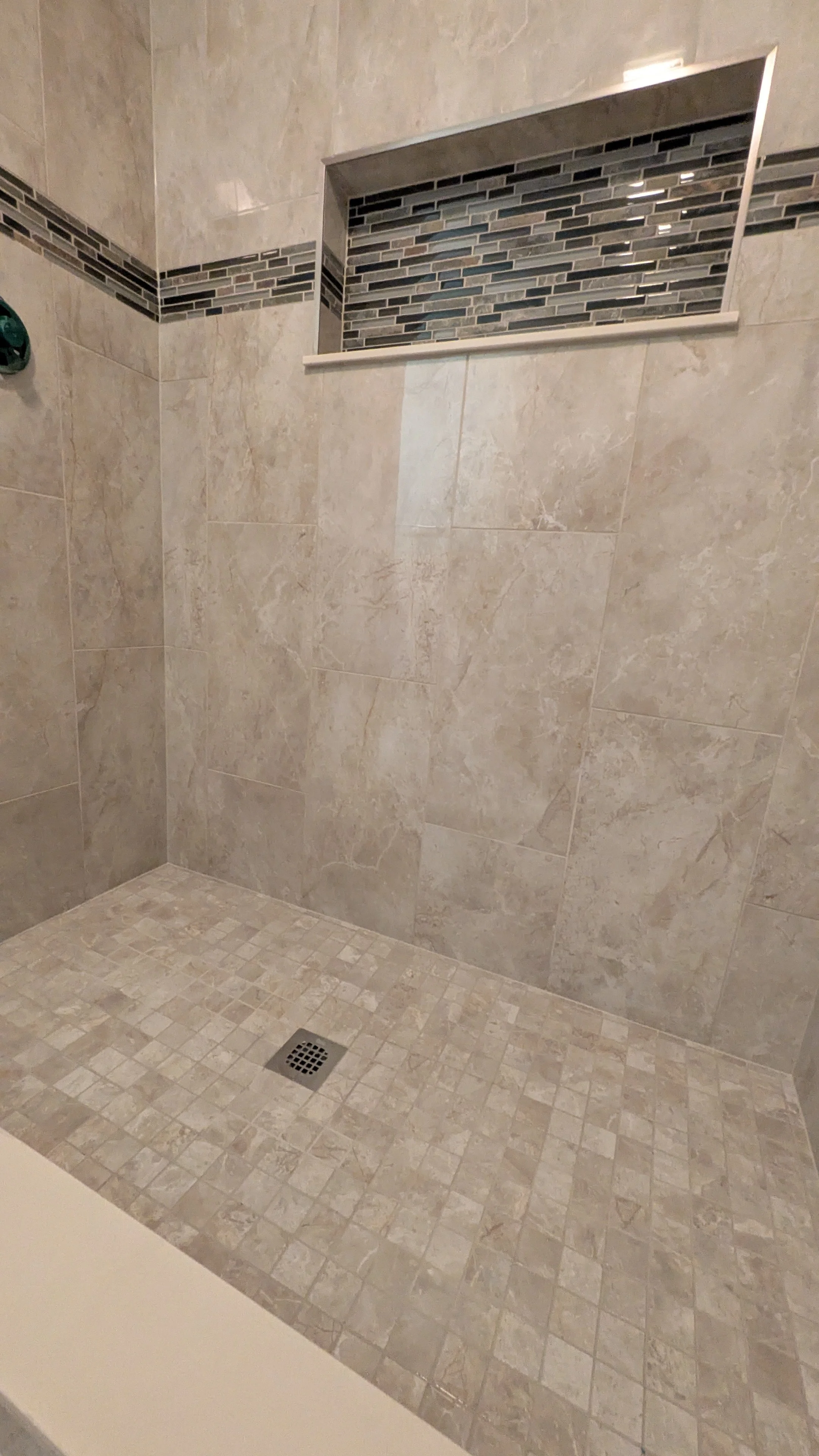 View of a tiled shower with beige large wall tiles, a mosaic accent strip, and a small niche with mosaic tiles in a recessed wall.