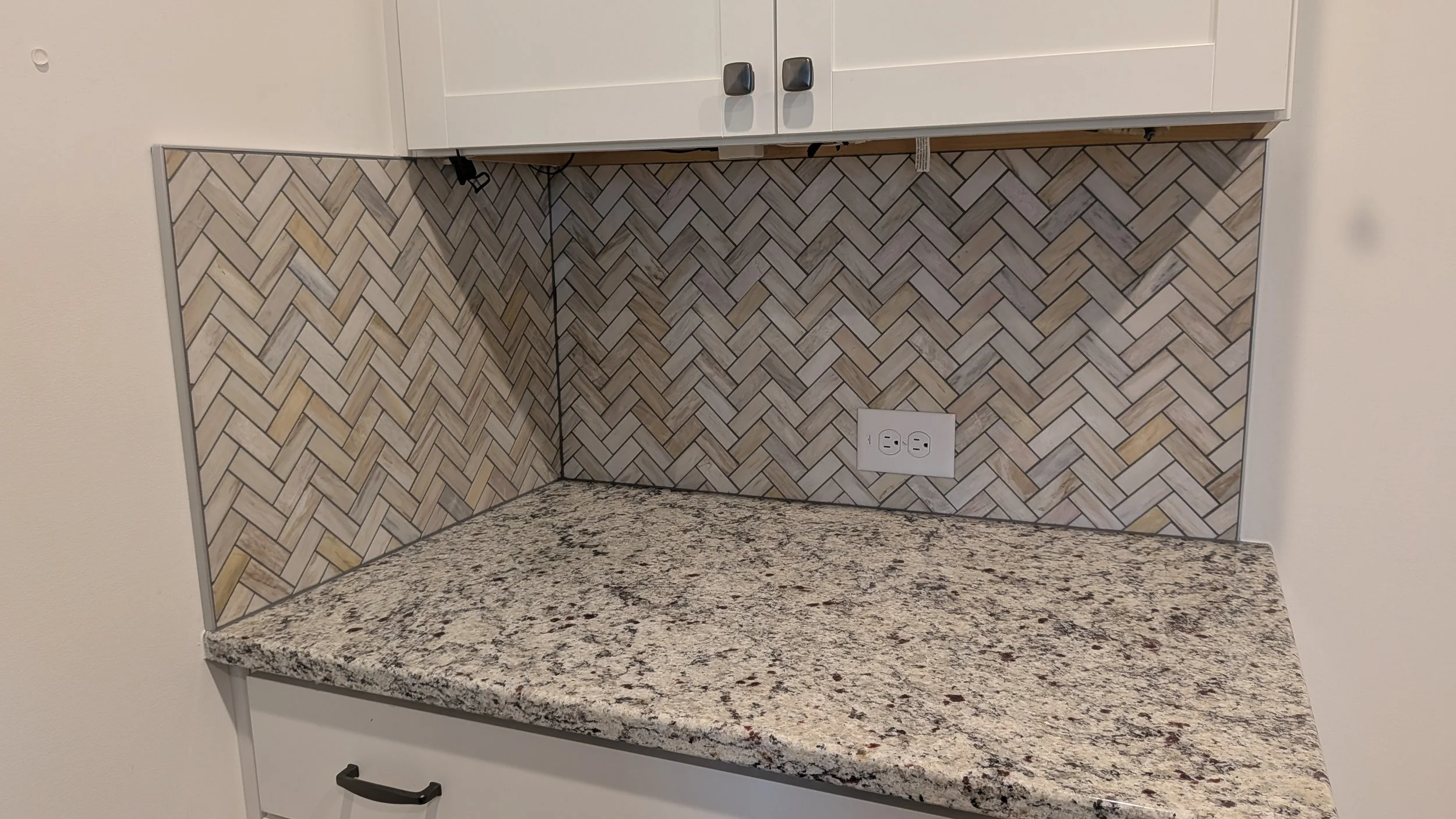 Kitchen countertop with a granite surface, white cabinets, herringbone patterned tile backsplash, and electrical outlets.