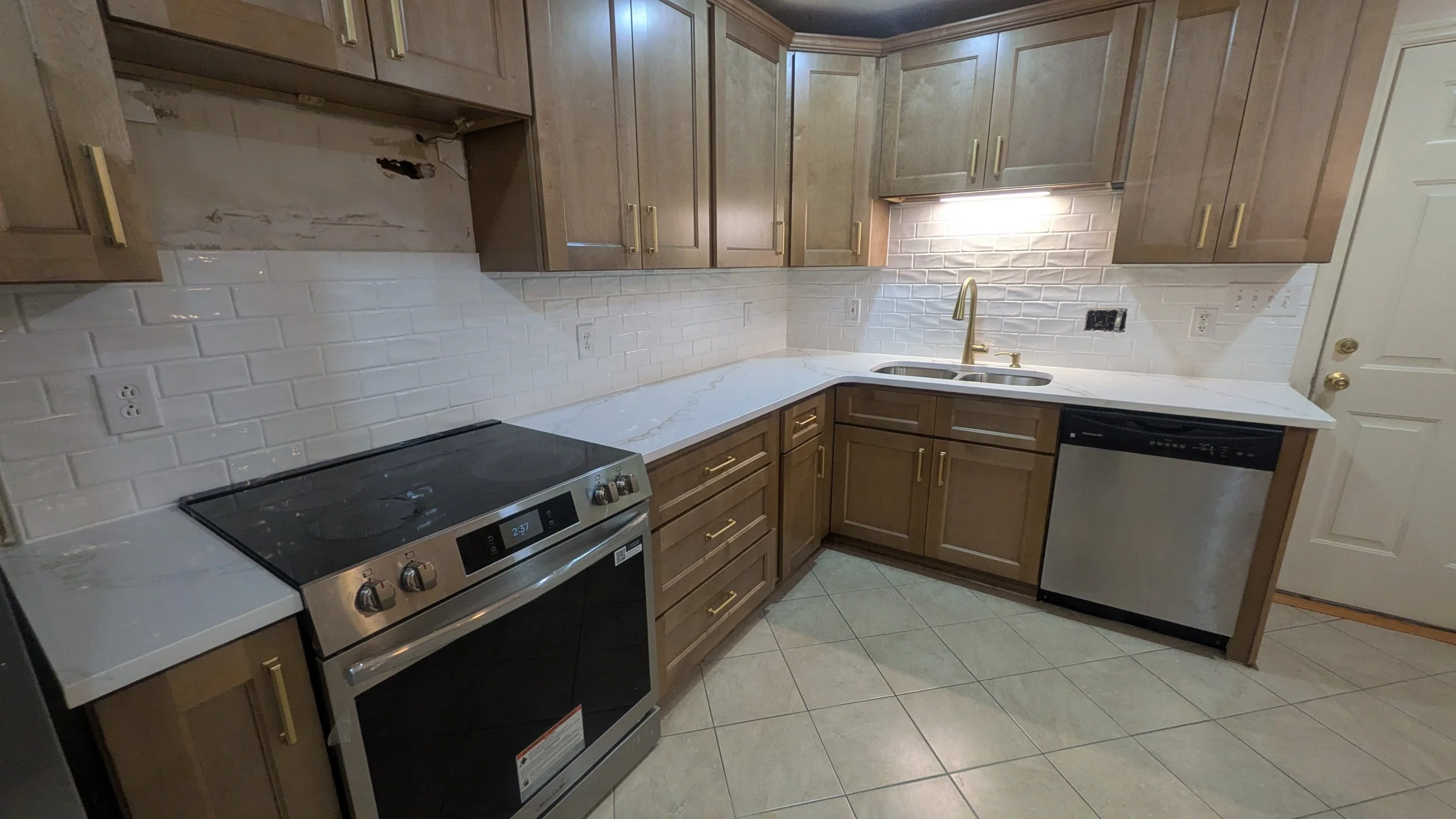 Empty kitchen with wooden cabinets, white marble countertops, white brick tile backsplash, stainless steel appliances including an oven and dishwasher, and a gold faucet over a double sink.