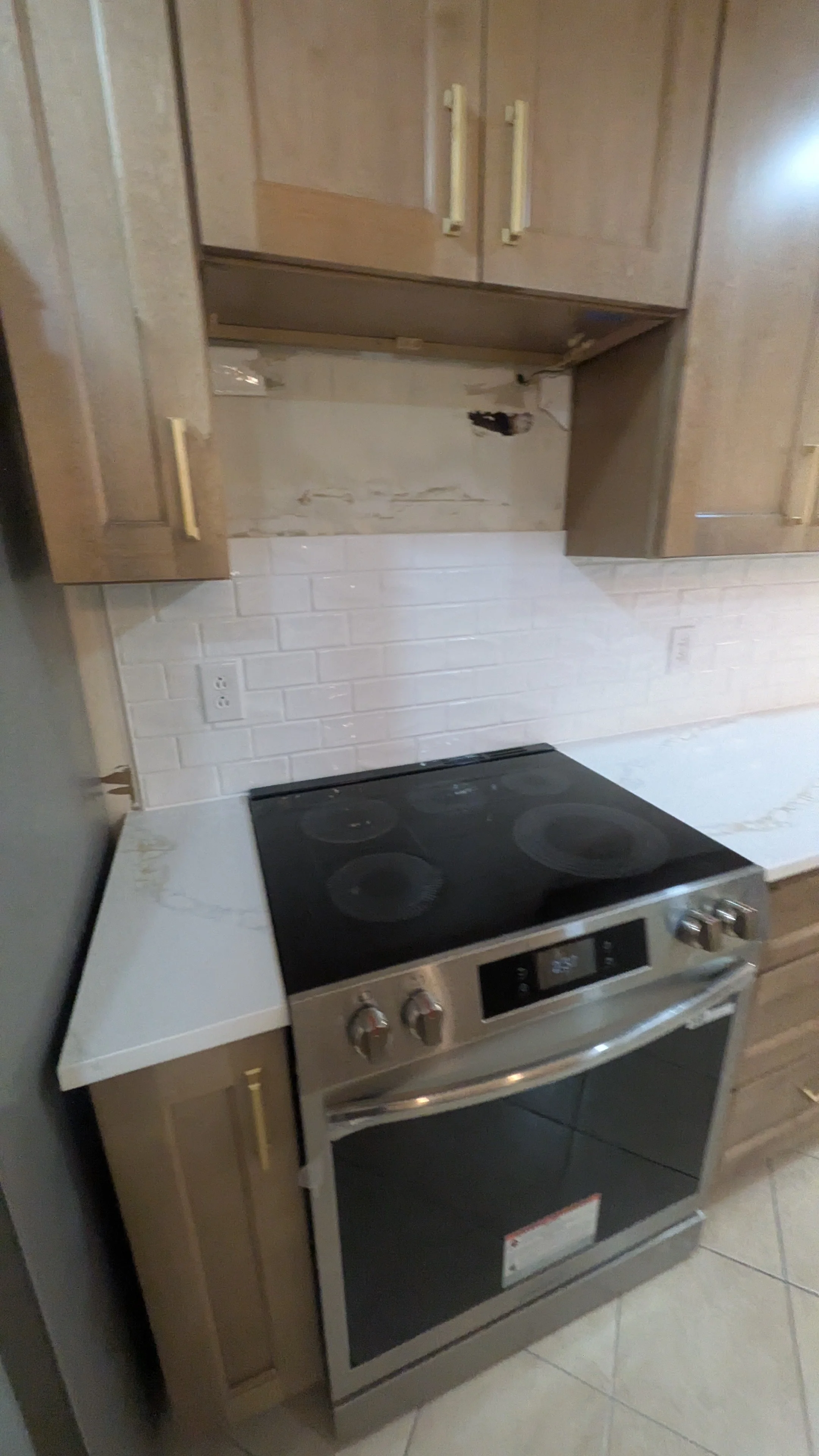 Kitchen with a black and silver stove, wooden cabinets, a white marble countertop, and a damaged backsplash area.