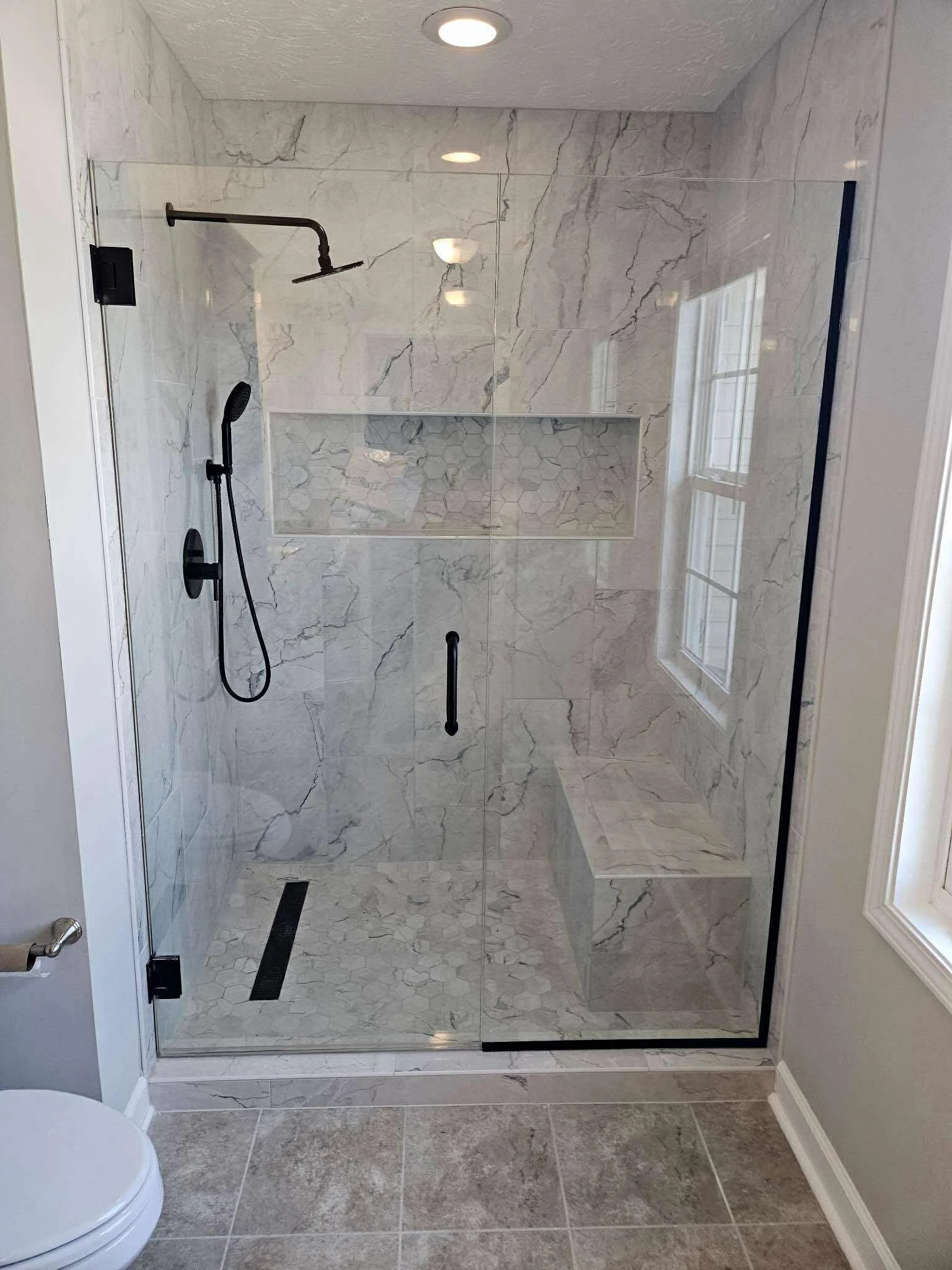 A modern walk-in shower with glass doors, marble tiles, a built-in bench, and two showerheads, one fixed and one handheld.