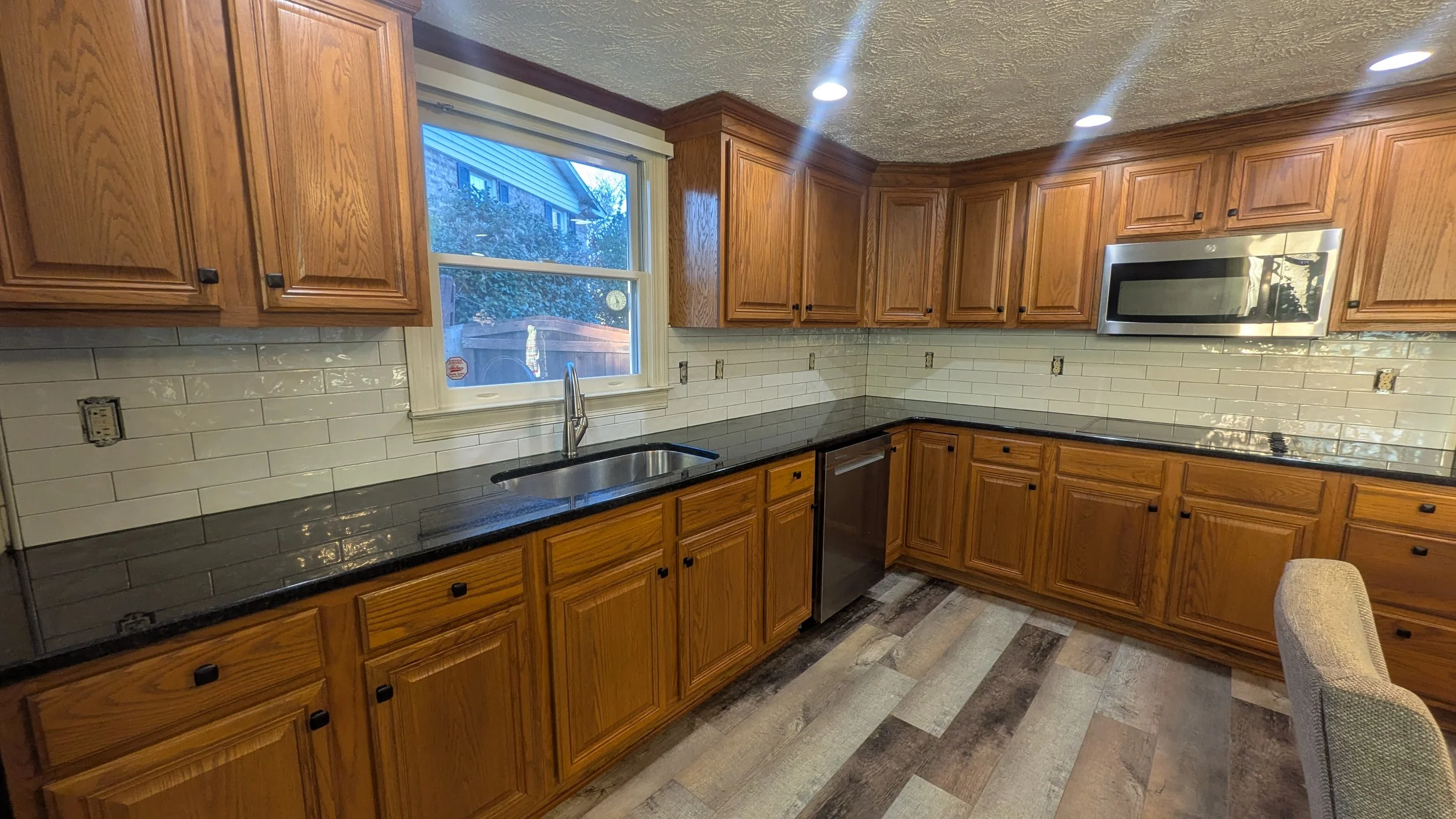 Kitchen with wooden cabinets, a window above a black countertop, white subway tile backsplash, a stainless steel sink, and built-in appliances, including a microwave.
