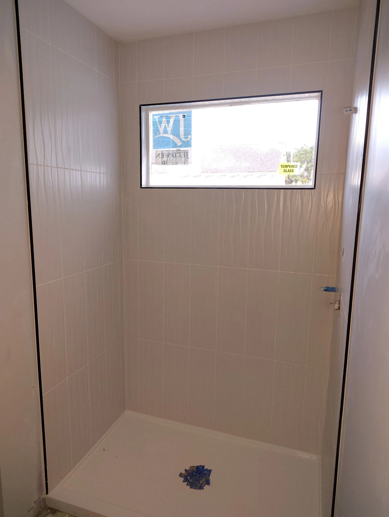 Empty shower with tiled walls, a rectangular window, and a drain at the bottom. The window has a sticker that says 'Tempered Glass'.