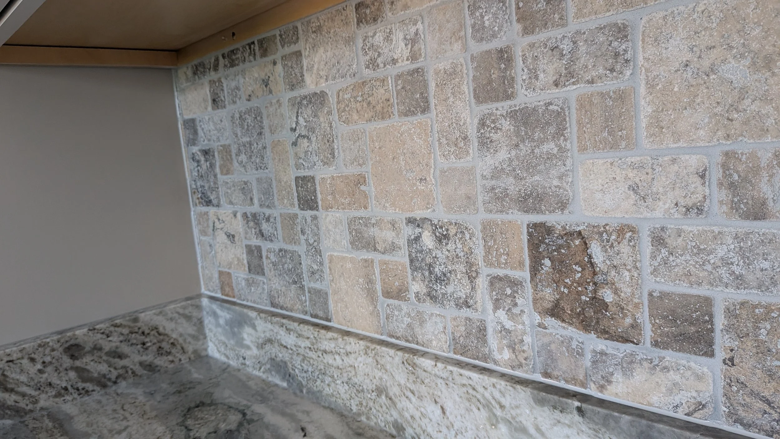 Close-up of beige and brown textured stone tile backsplash in a kitchen, with a granite countertop in the foreground.
