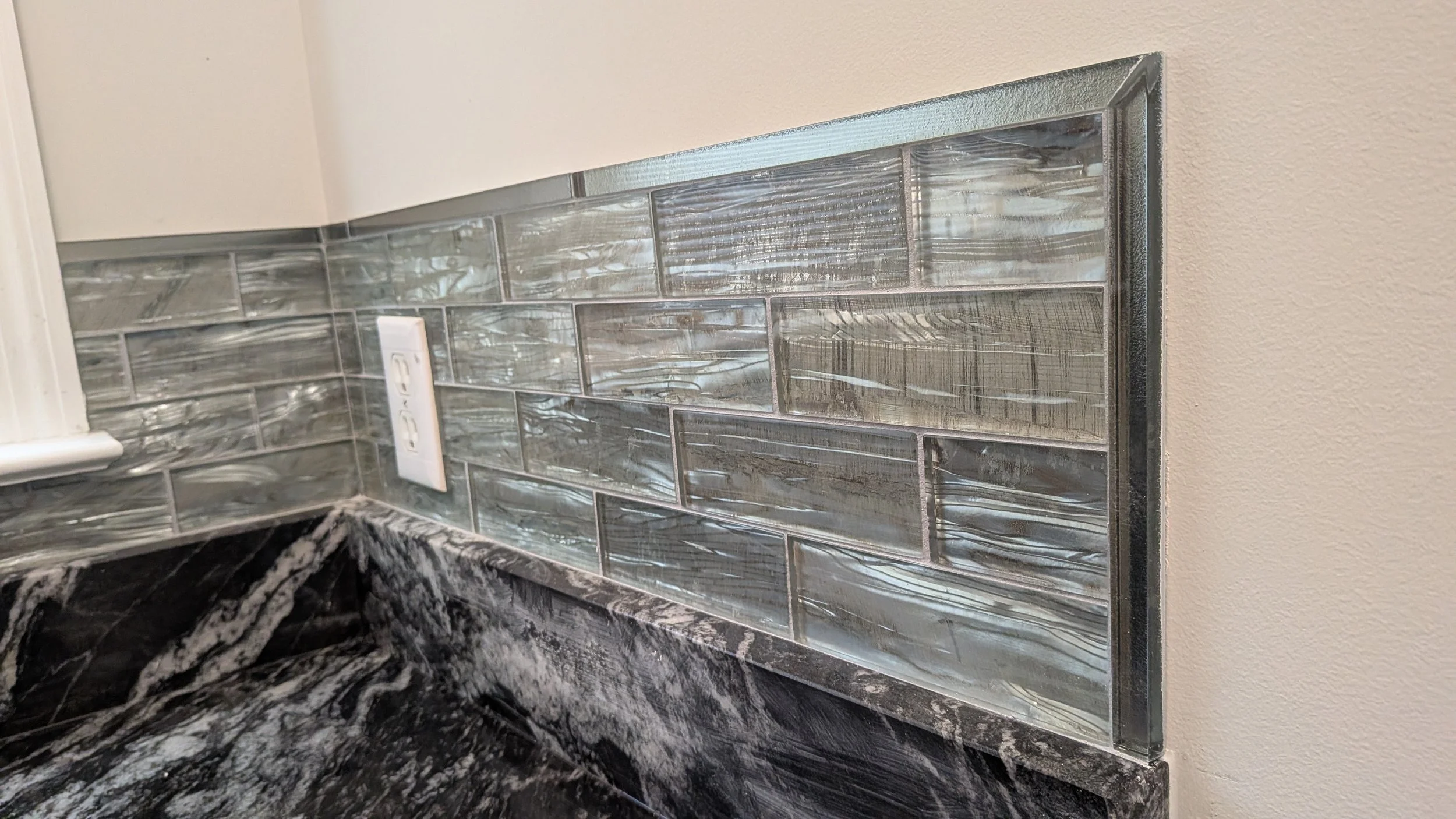 Kitchen backsplash with grey and iridescent glass subway tiles and marble countertop, including a white electrical outlet.