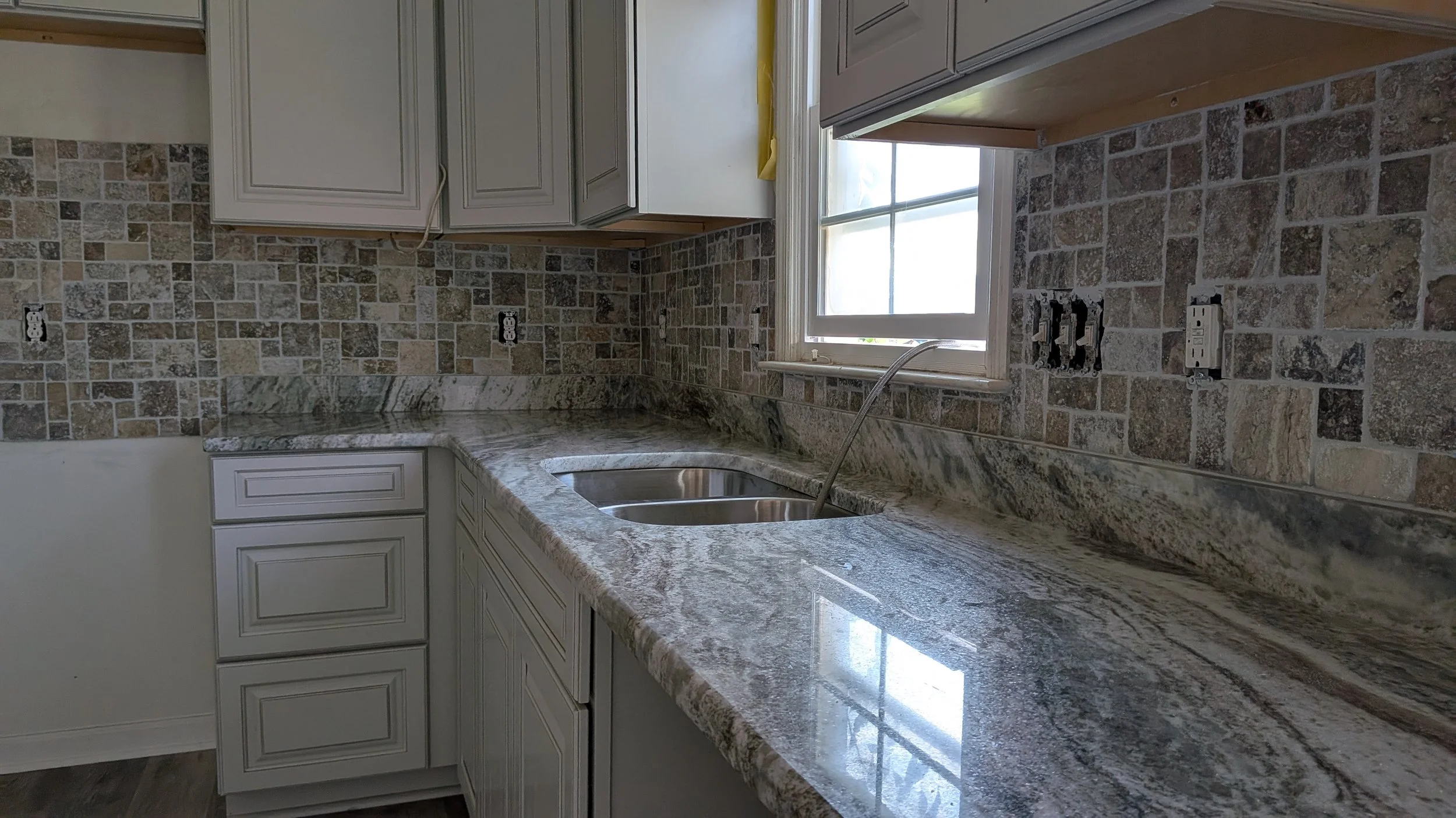 Kitchen with gray cabinets, granite countertops, and a brick-style backsplash. There is a double sink under a window, with outlets and switches on the wall.
