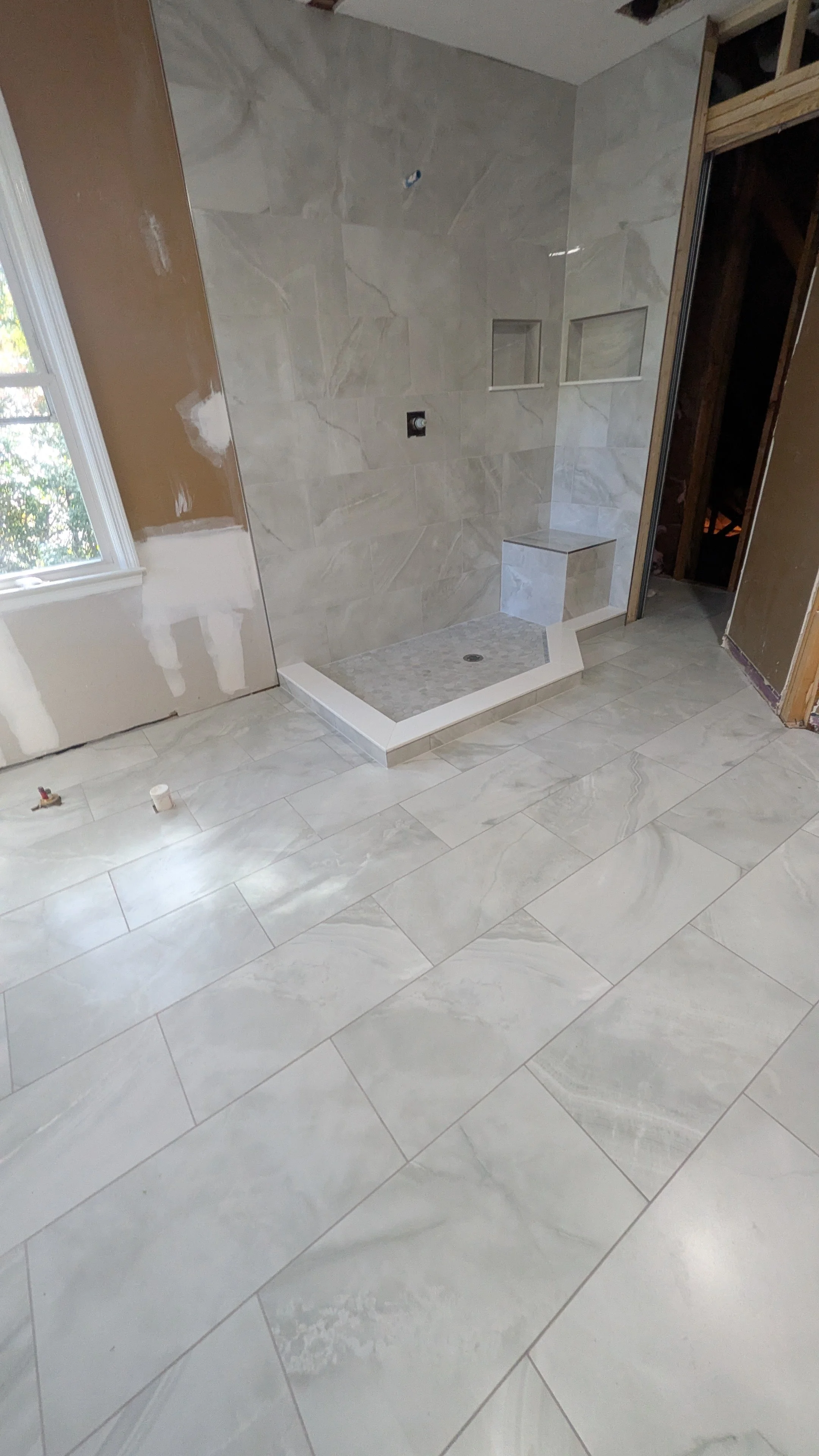 Interior of a bathroom under construction, showing a tiled shower area with built-in shelves and a small bench, with unfinished walls and a window on the left side.