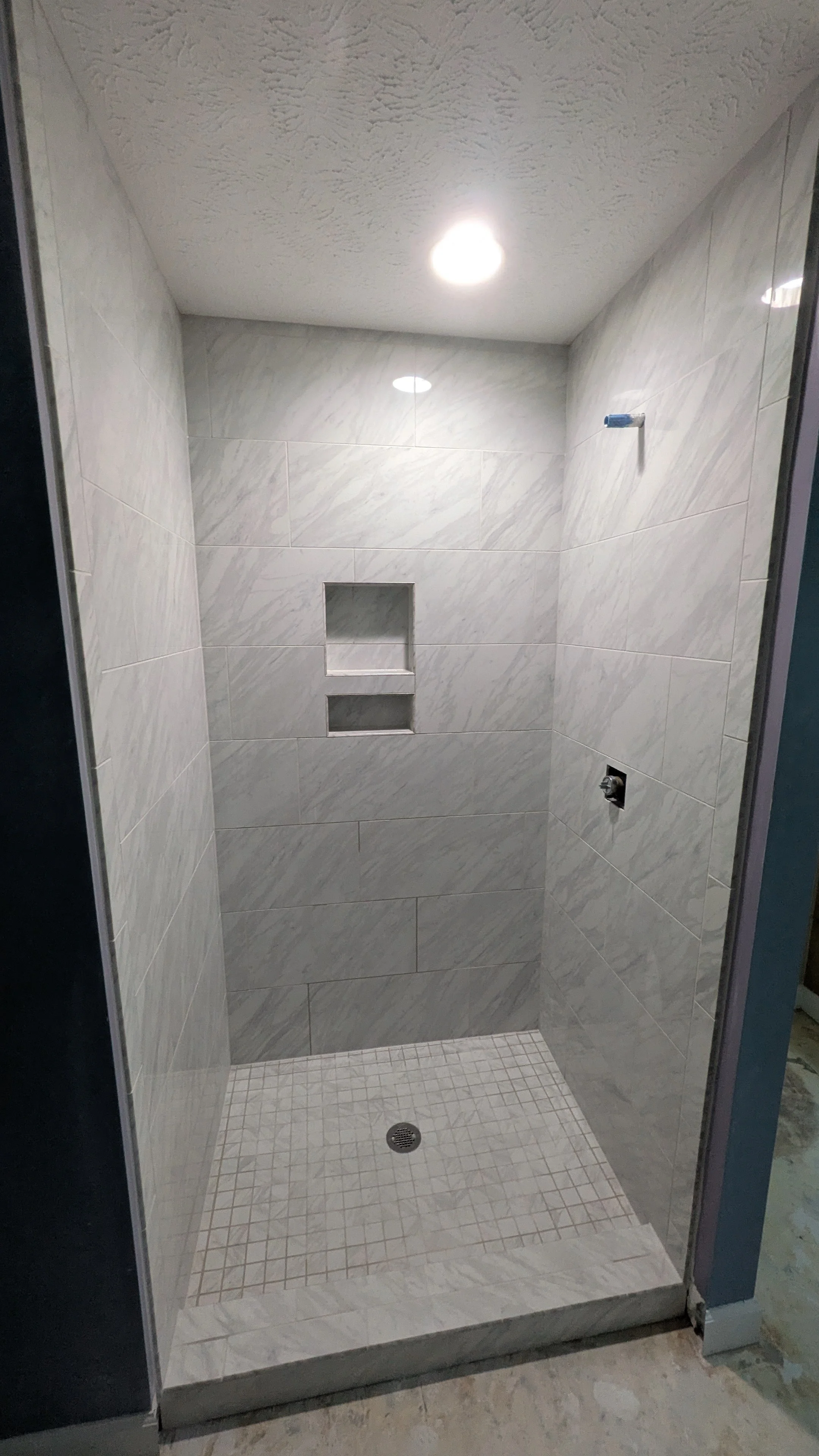 A newly constructed walk-in shower with gray marble tiles, built-in shelves, and a ceiling light fixture.