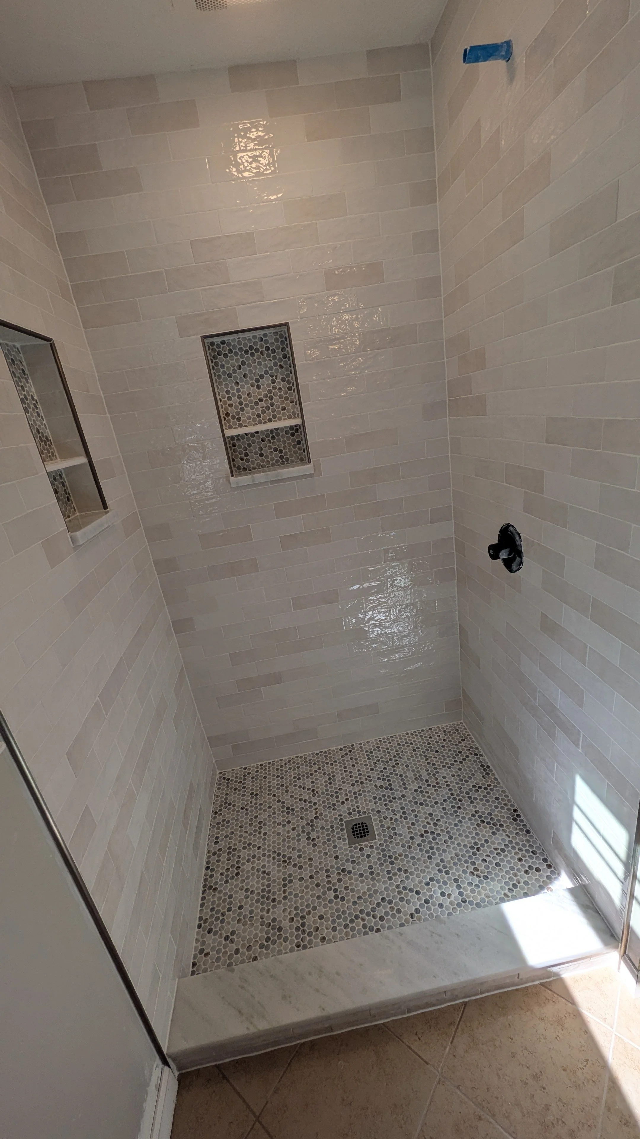 Empty tiled shower with built-in shelves, a drain, and a small window, in a bathroom with beige tiles.