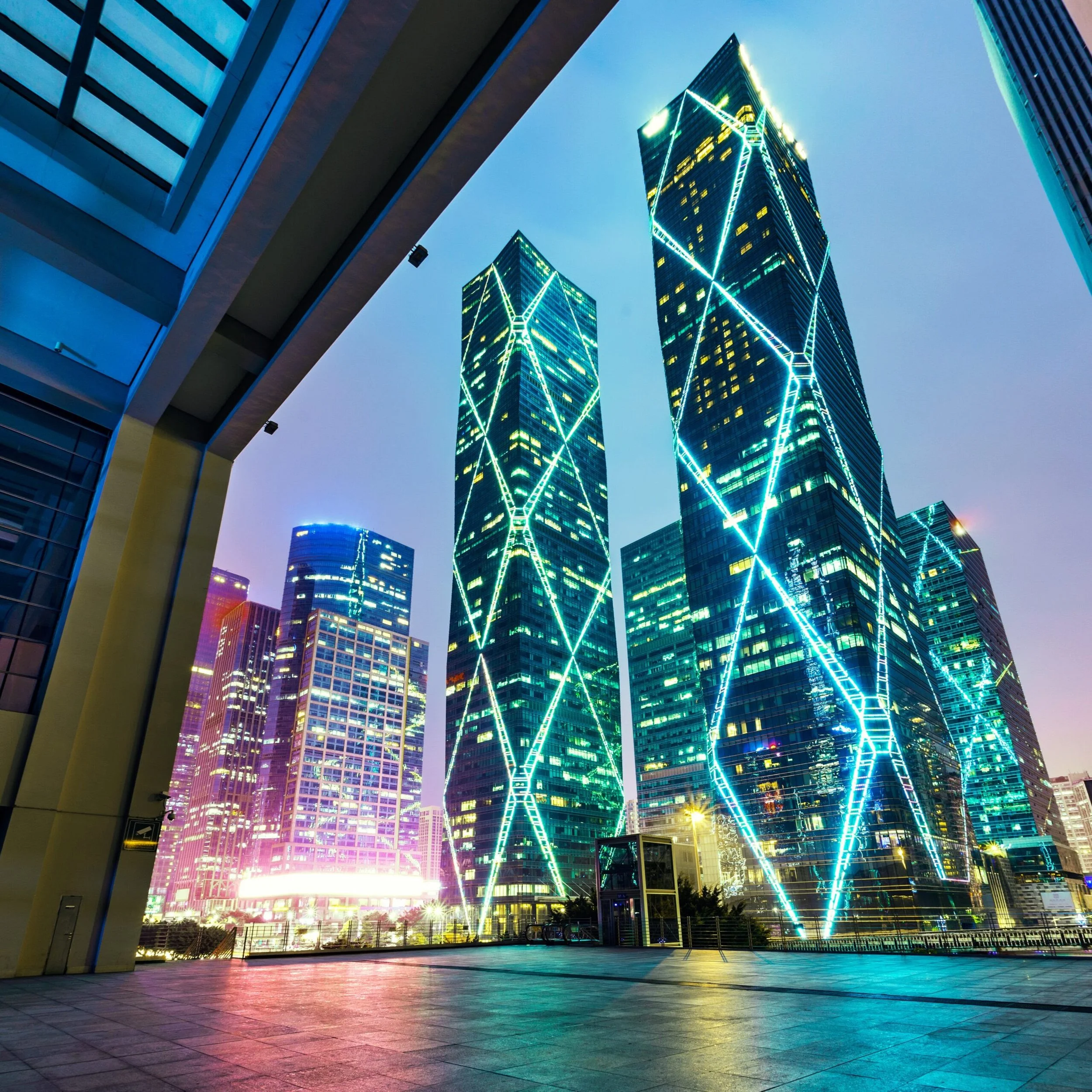 Nighttime cityscape with illuminated skyscrapers featuring neon light patterns, viewed from a low angle with a building arch framing the scene.