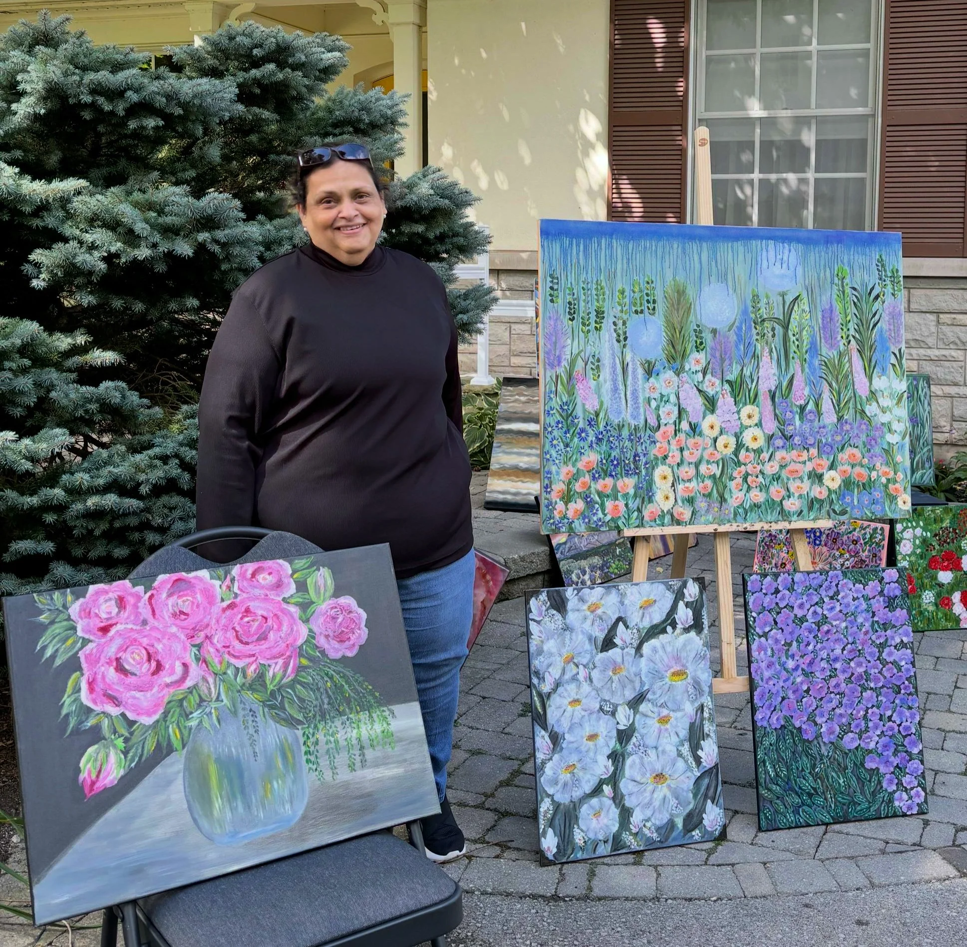 A woman stands outdoors next to her floral paintings displayed on a chair and easels, with a large evergreen tree and a house in the background.