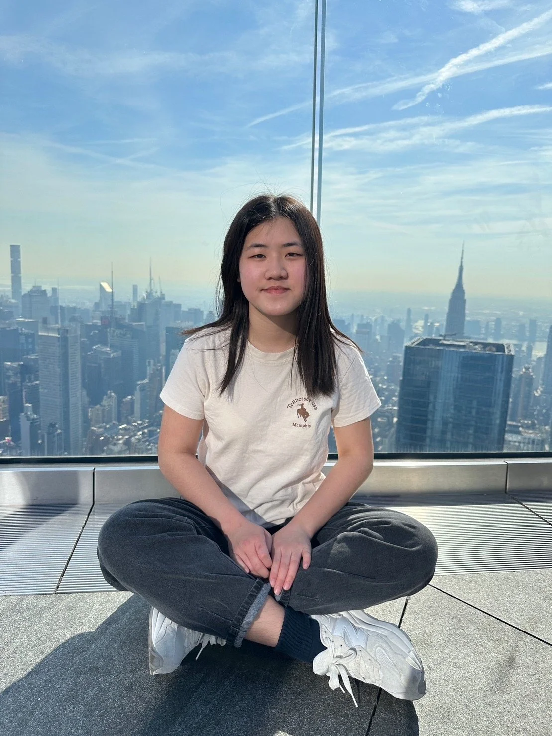 Young woman sitting cross-legged on a rooftop in front of a large glass window, with the New York City skyline, including the Empire State Building, in the background on a clear day.