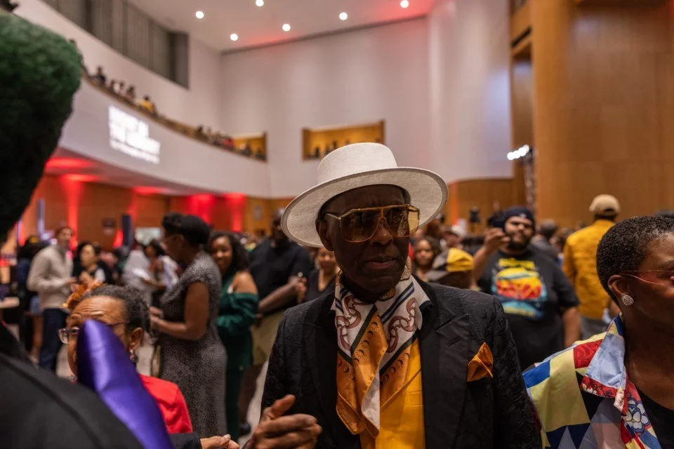 A man wearing a white hat, large sunglasses, a scarf, and a suit at a crowded indoor event