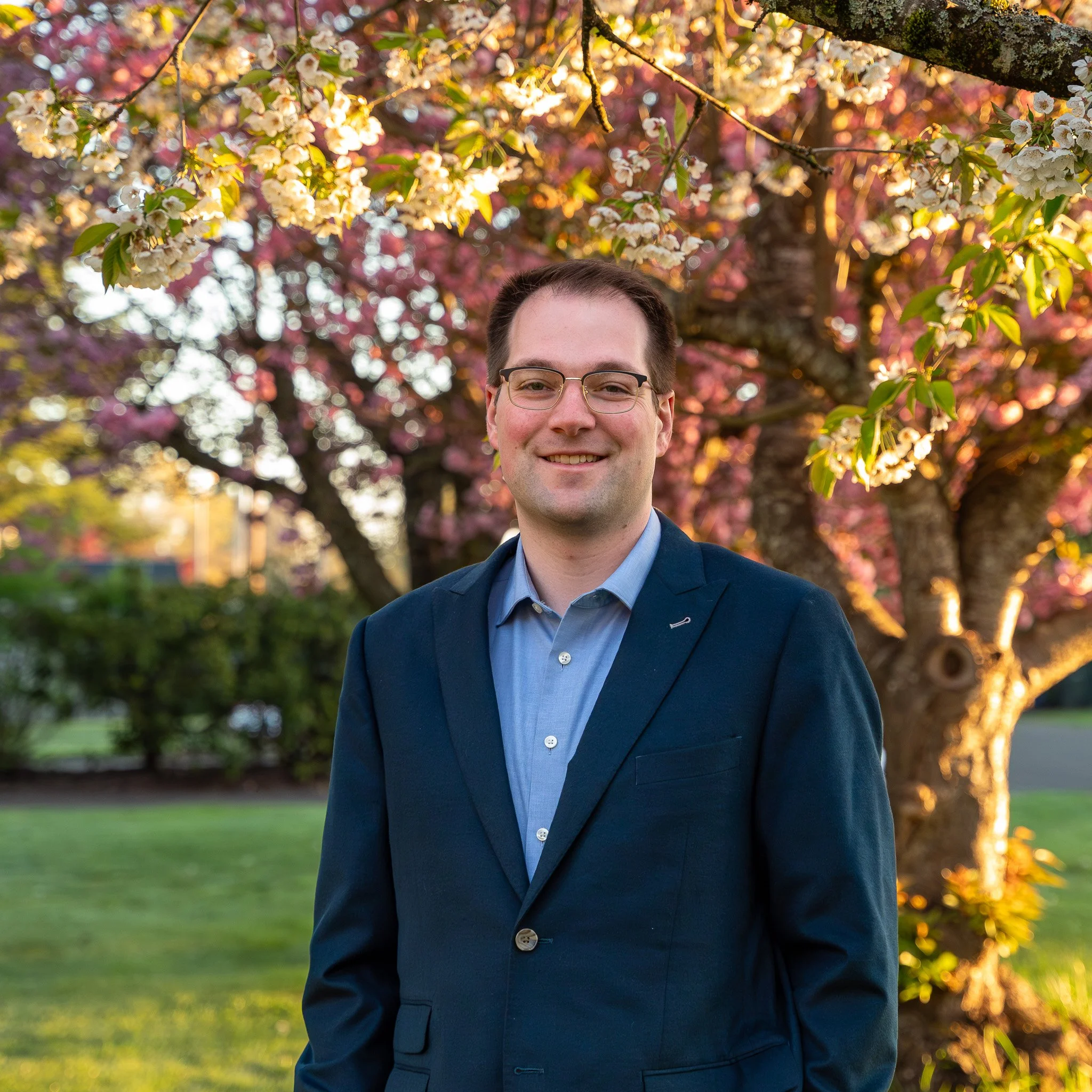 Matt standing in an orchard with green leaves behind him, wearing glasses and a light blue button-down shirt, smiling warmly at the camera.