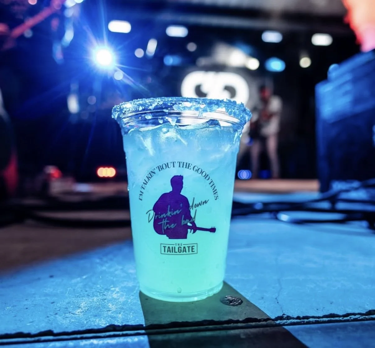 A blue cocktail with ice in a plastic cup, decorated with a rim of salt, sitting on a table in a dimly lit bar or event space, with a blurred stage and people in the background.