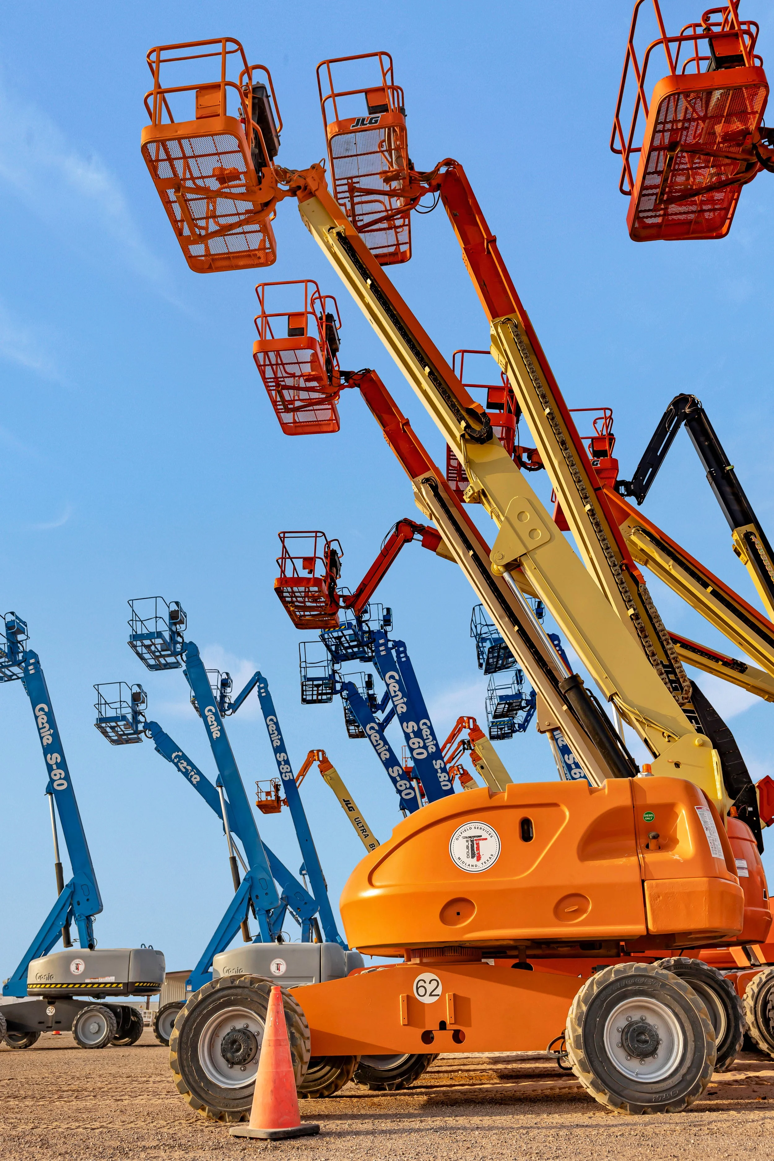 Multiple mobile boom lifts in orange and blue, parked on a dirt lot under a clear blue sky, with orange traffic cone in the foreground.