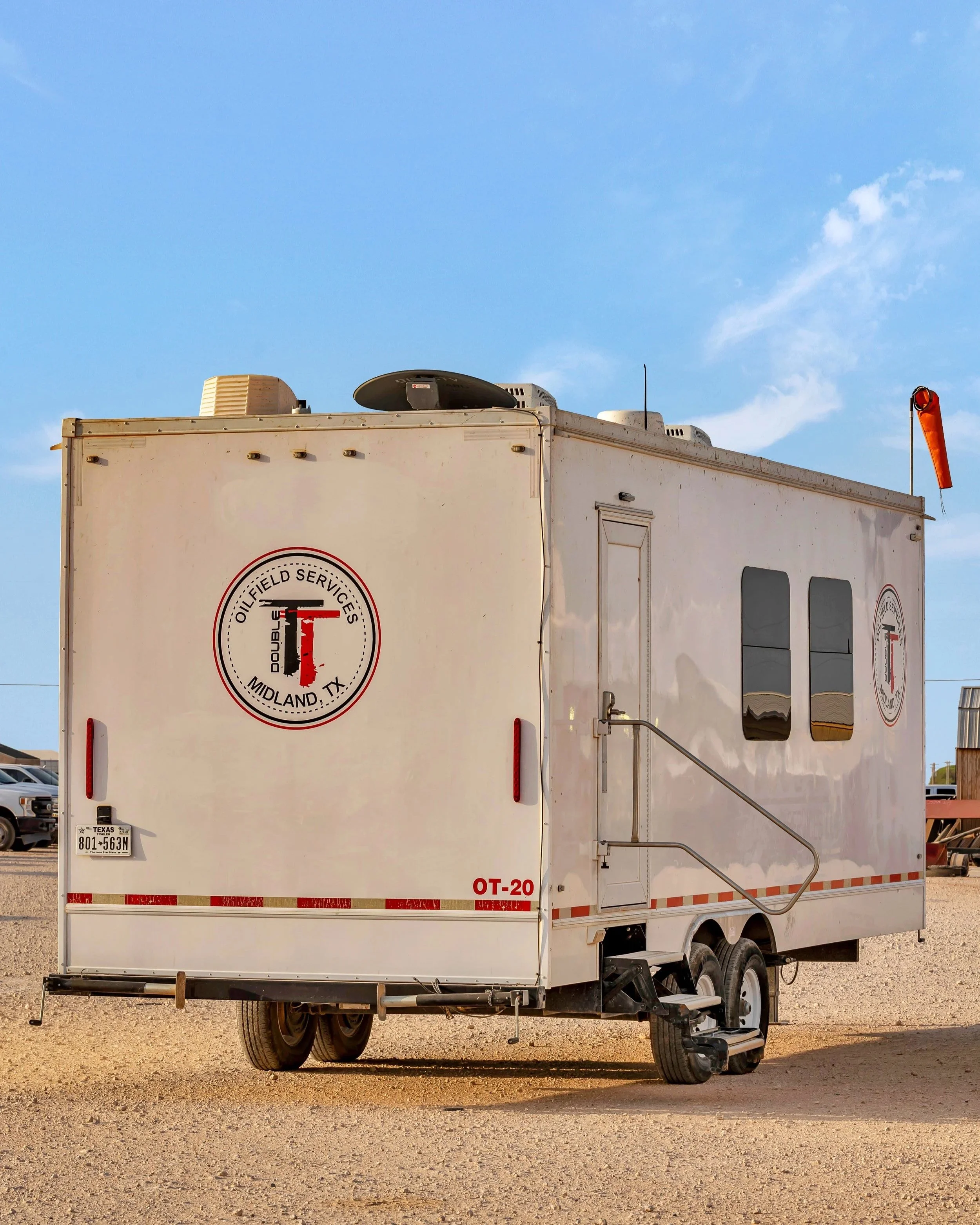 Mobile oilfield services trailer with Midland, Texas logo, two small windows, a door with a handrail, and a Texas license plate, set against a blue sky with few clouds.