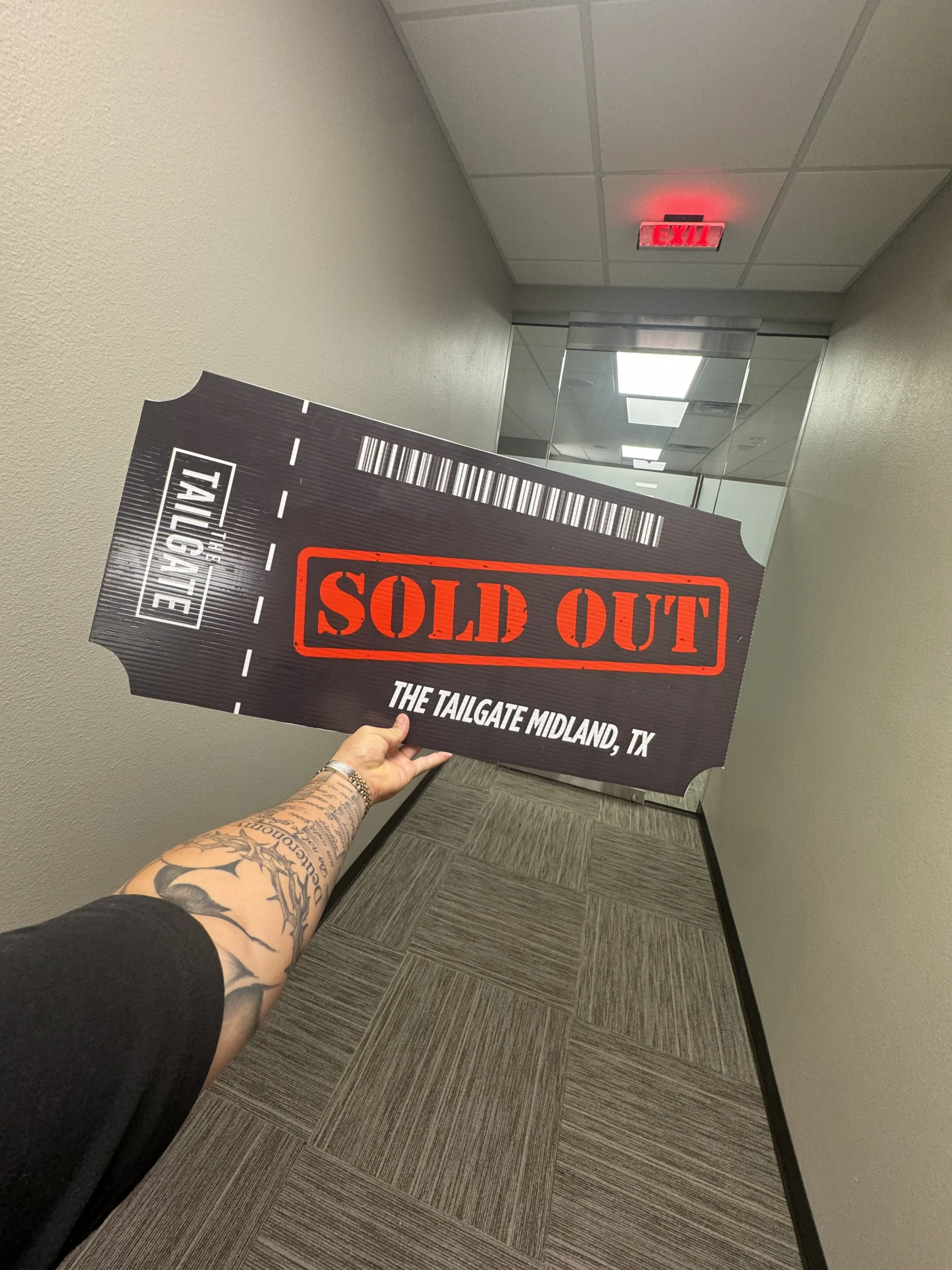A person holding a large black sign shaped like a ticket that reads 'SOLD OUT' in red letters, with the location 'THE TAILGATE MIDLAND, TX' at the bottom, in a hallway with a mirror and an illuminated exit sign.
