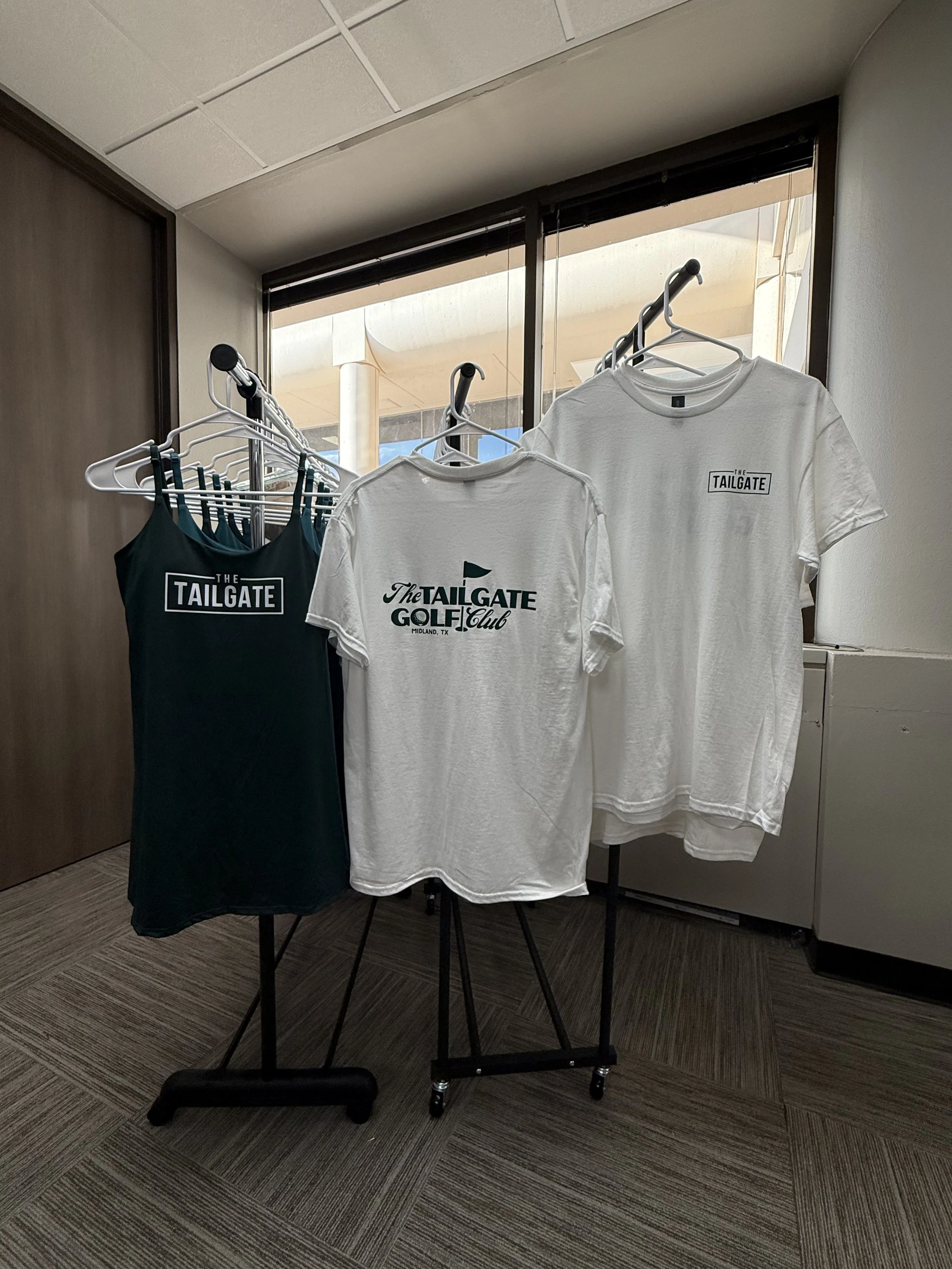 Display of tailgate-themed merchandise including a teal dress and T-shirts with 'The Tailgate' logo, on a rack and table in an indoor setting.