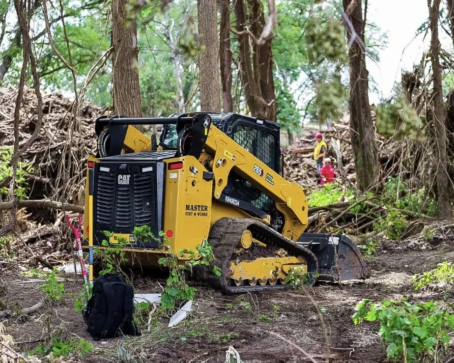A yellow Cat mini bulldozer clearing a fallen tree in a forest, with two people in the background.