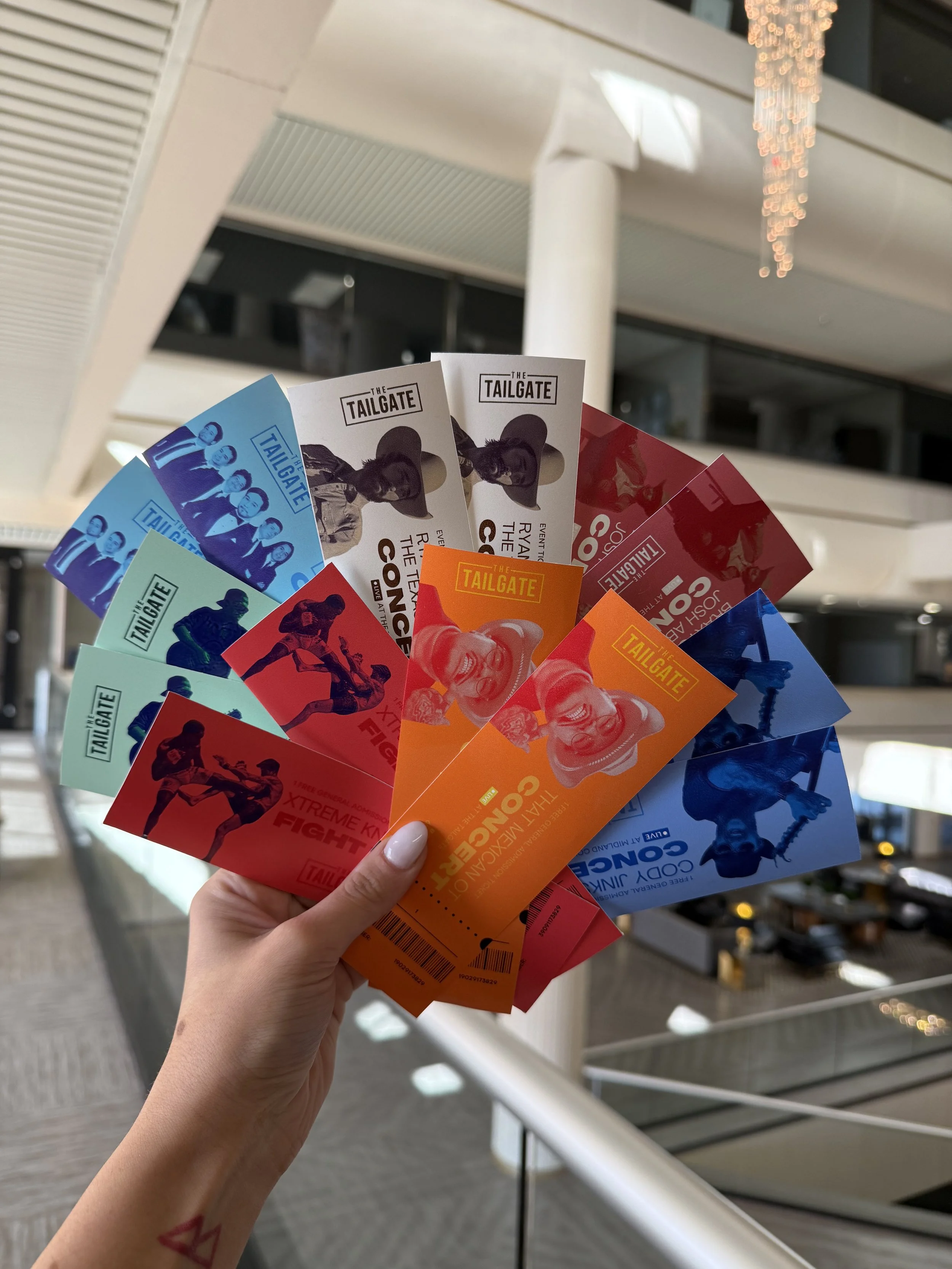 A person's hand holding multiple colorful tickets for The Tailgate event, with various images and text, inside a modern building.
